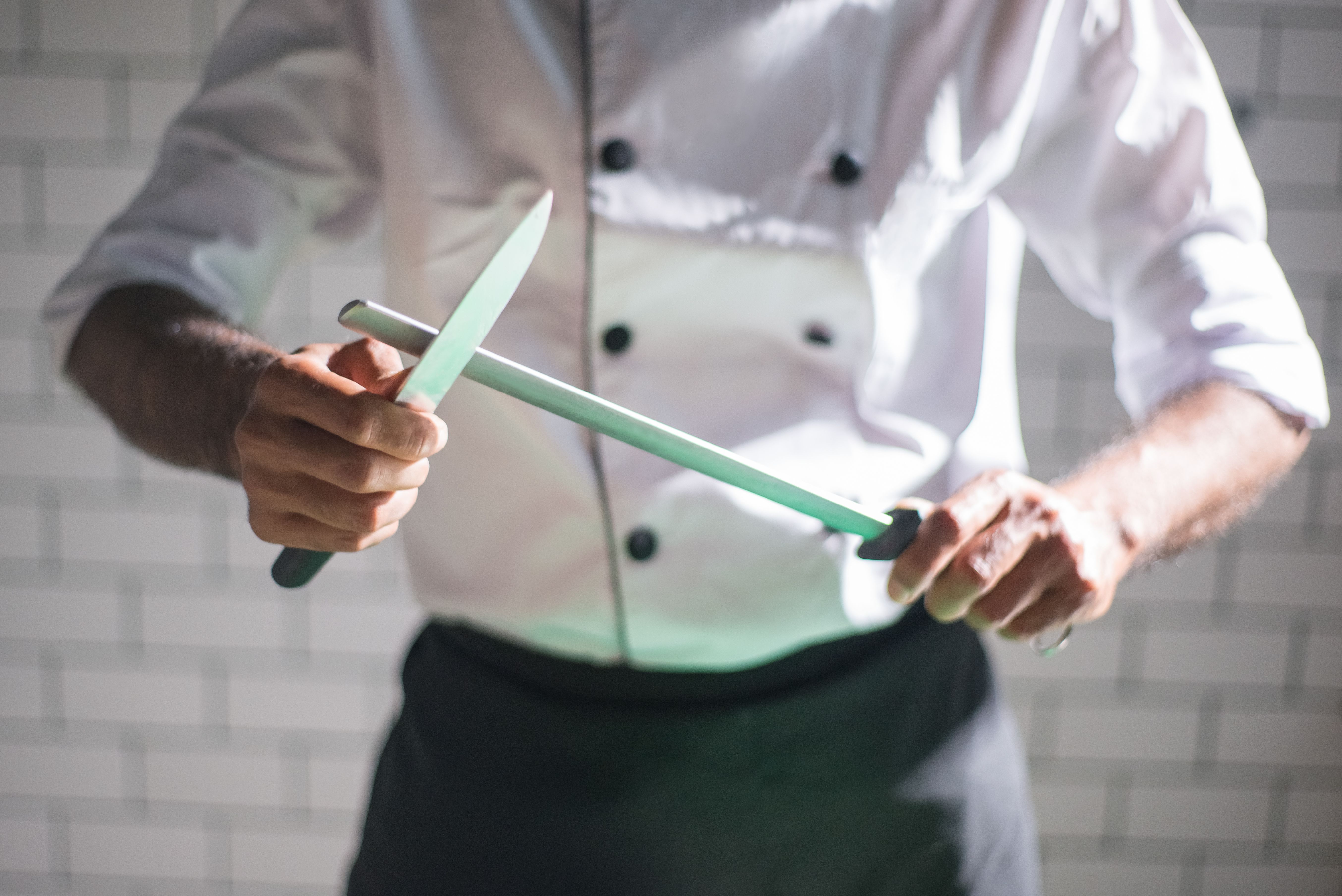A chef in a white uniform sharpening a large kitchen knife using a honing steel rod.
