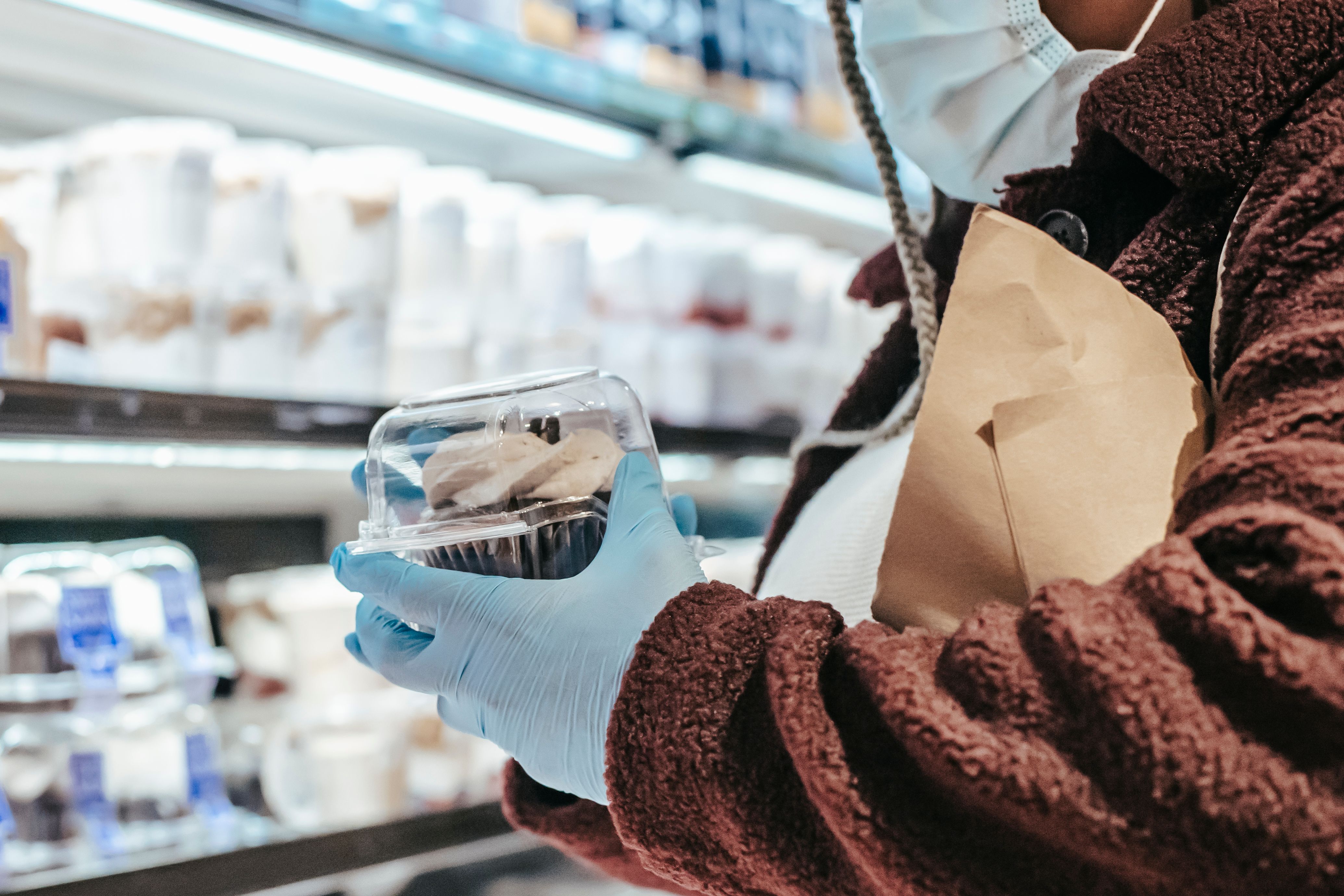 A shopper wearing a face mask and blue protective gloves inspecting a packaged cupcake in a store.