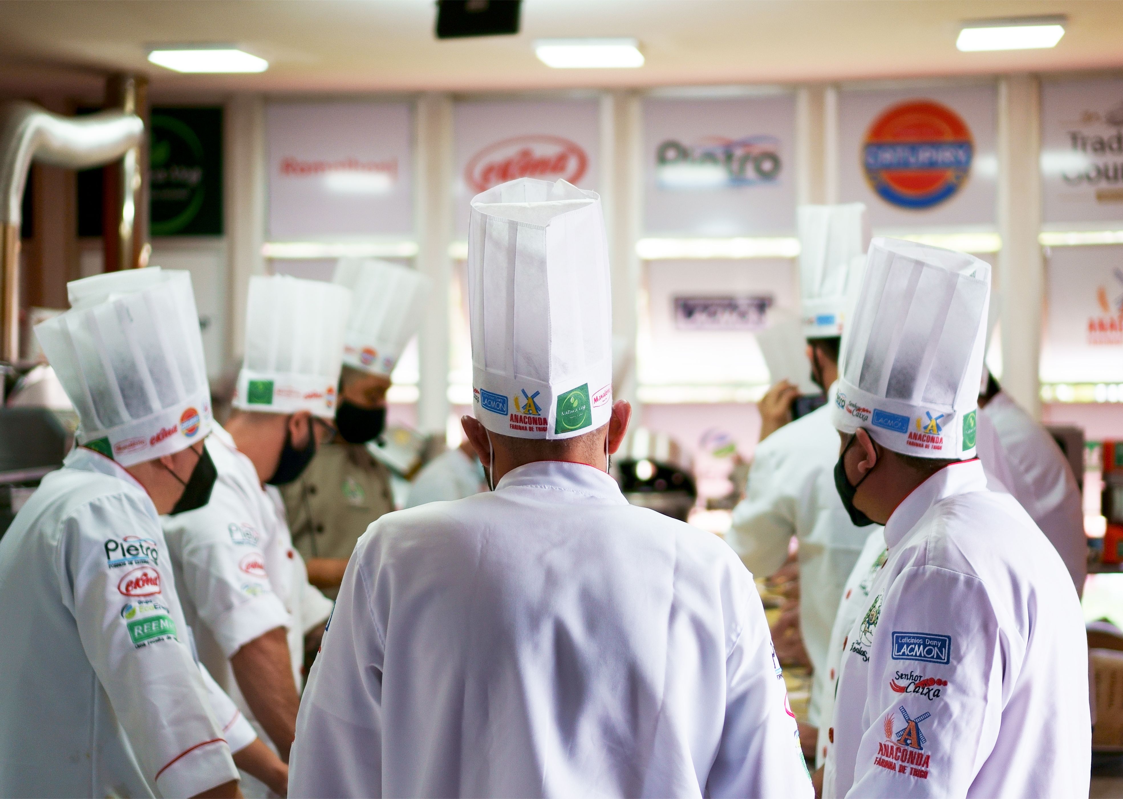 A rear view of professional chefs wearing tall white chef hats and branded uniforms gathered in a commercial kitchen.