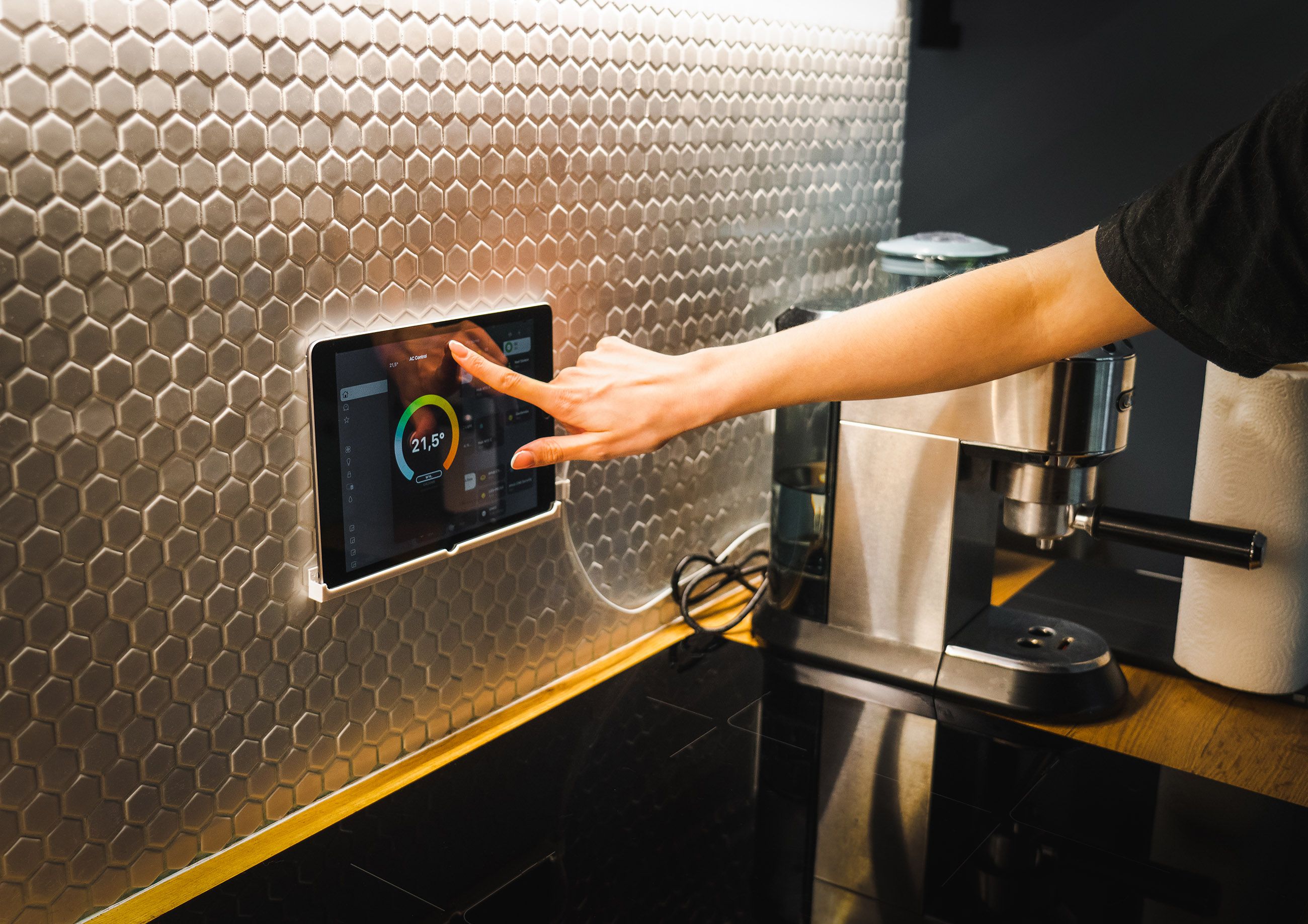 A person using a finger to adjust temperature settings on a smart wall-mounted digital display screen in a kitchen next to a coffee machine.