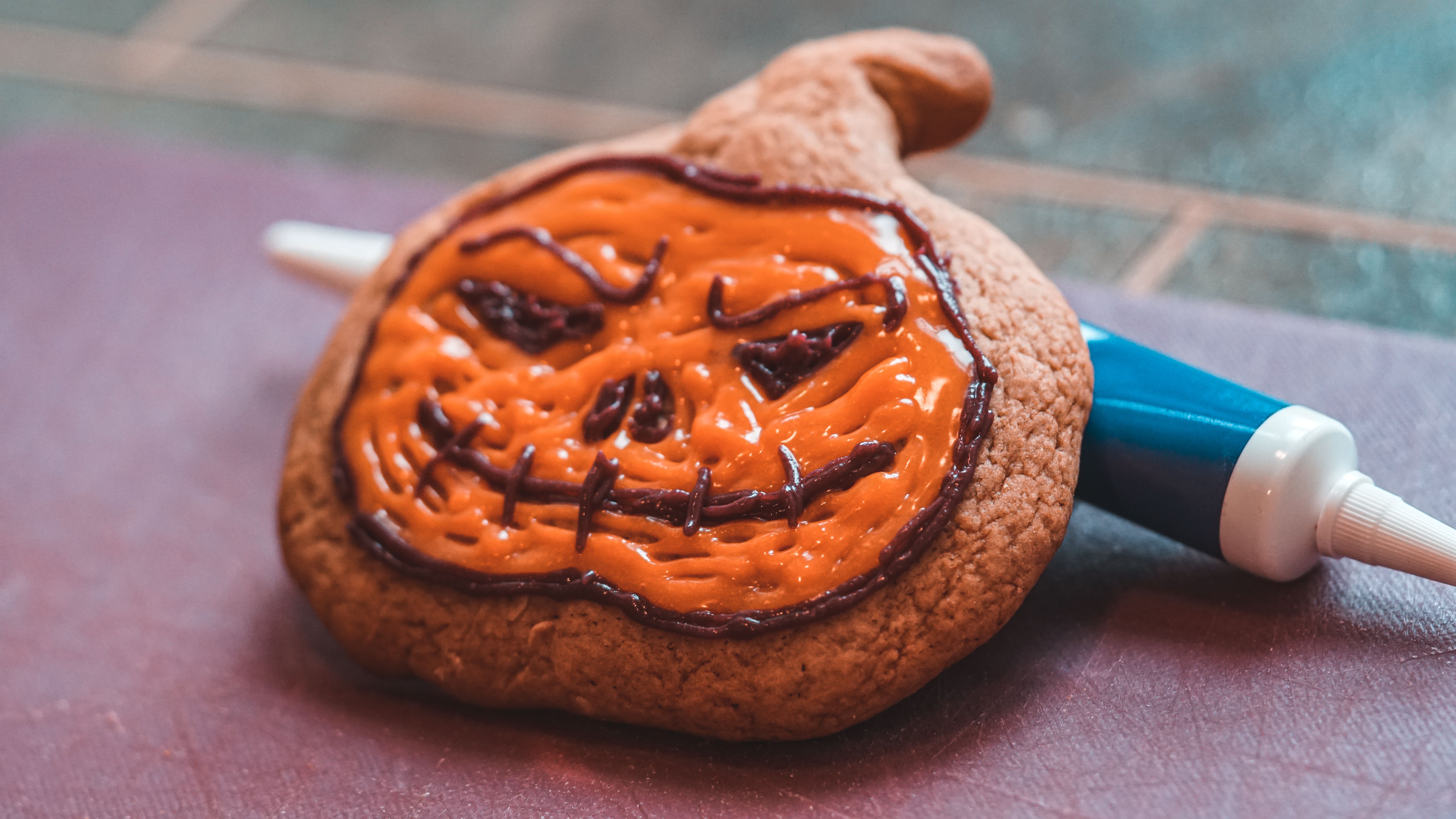 A pumpkin-shaped cookie decorated with orange and dark icing resting on a surface next to an icing tube