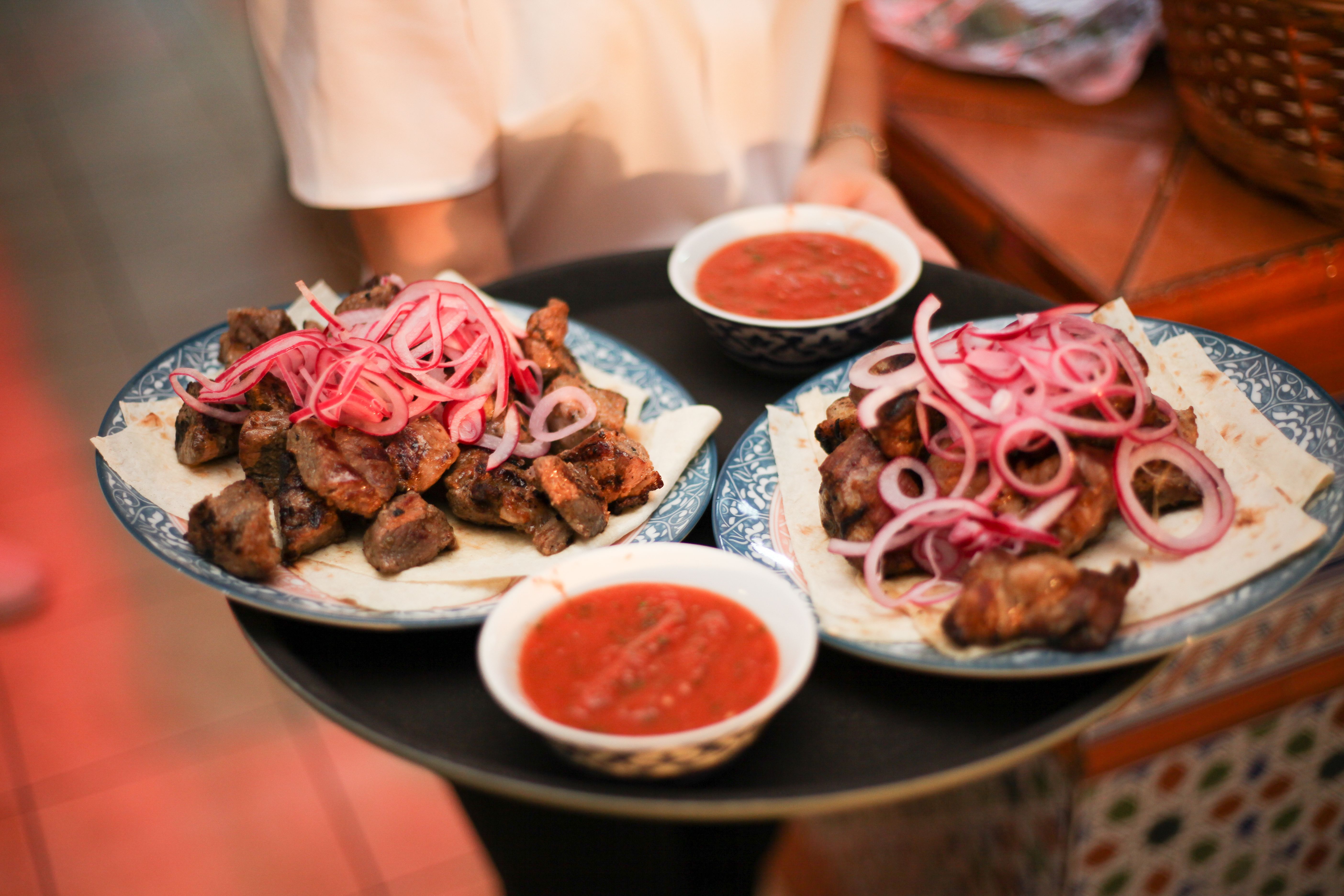 A server holding a tray with two plates of grilled meat topped with sliced red onions, accompanied by two small bowls of red salsa sauce.