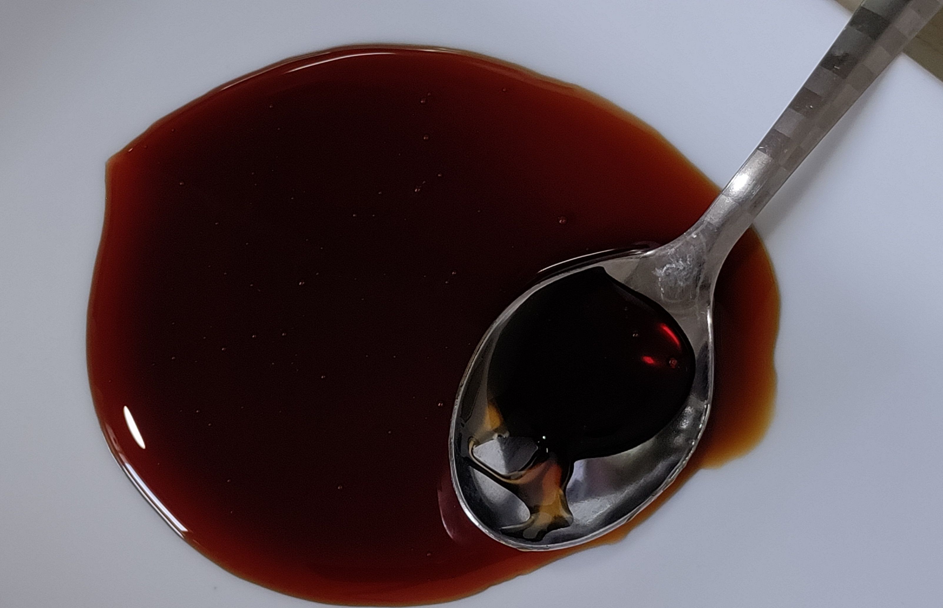 A top-down close-up view of a metal spoon dipping into a pool of dark reddish-brown liquid or syrup on a white plate.