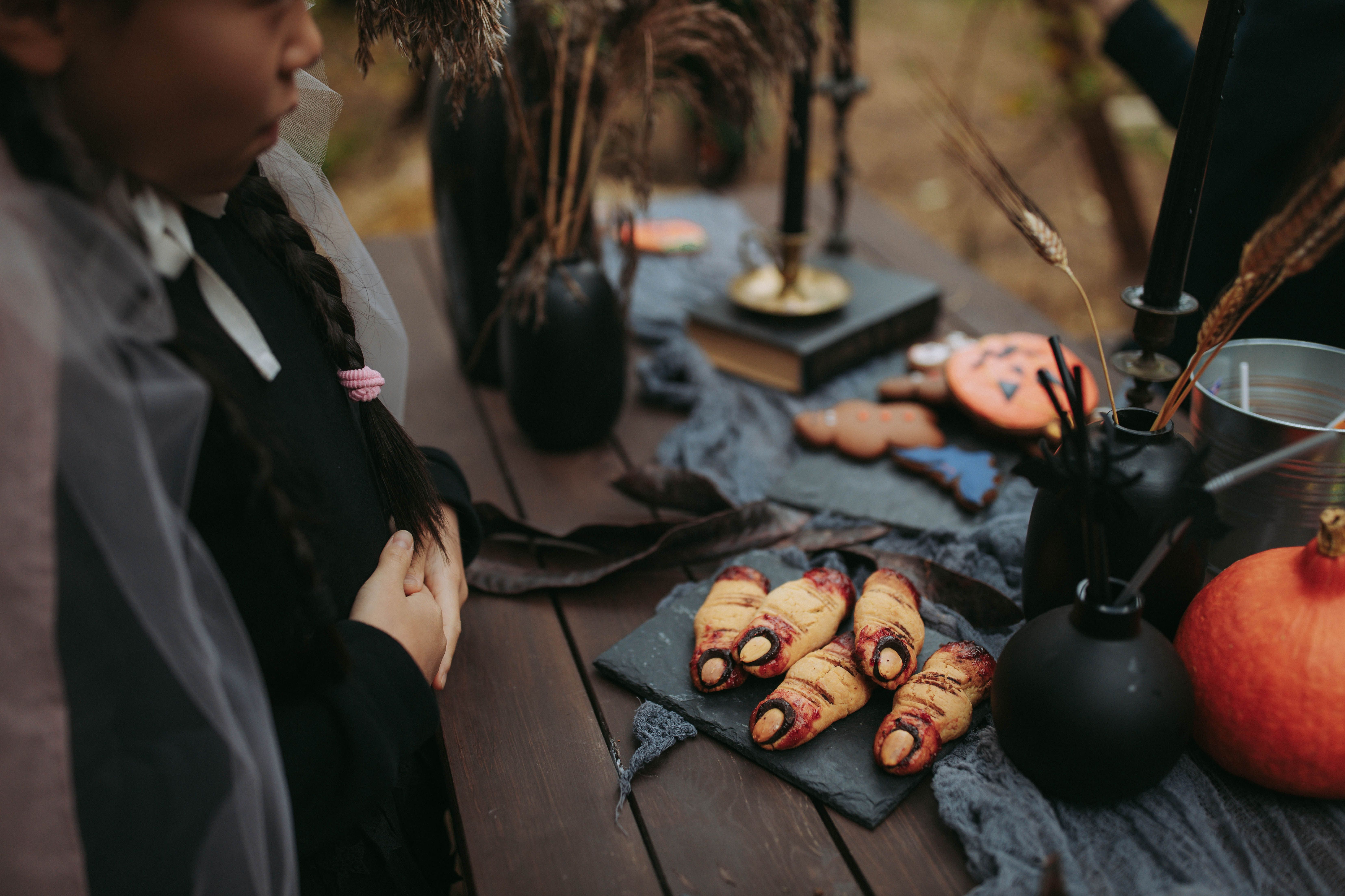 A person standing by a Halloween-themed table set with witch-themed decorations, pumpkins, and baked cookies shaped like severed, bloody fingers.