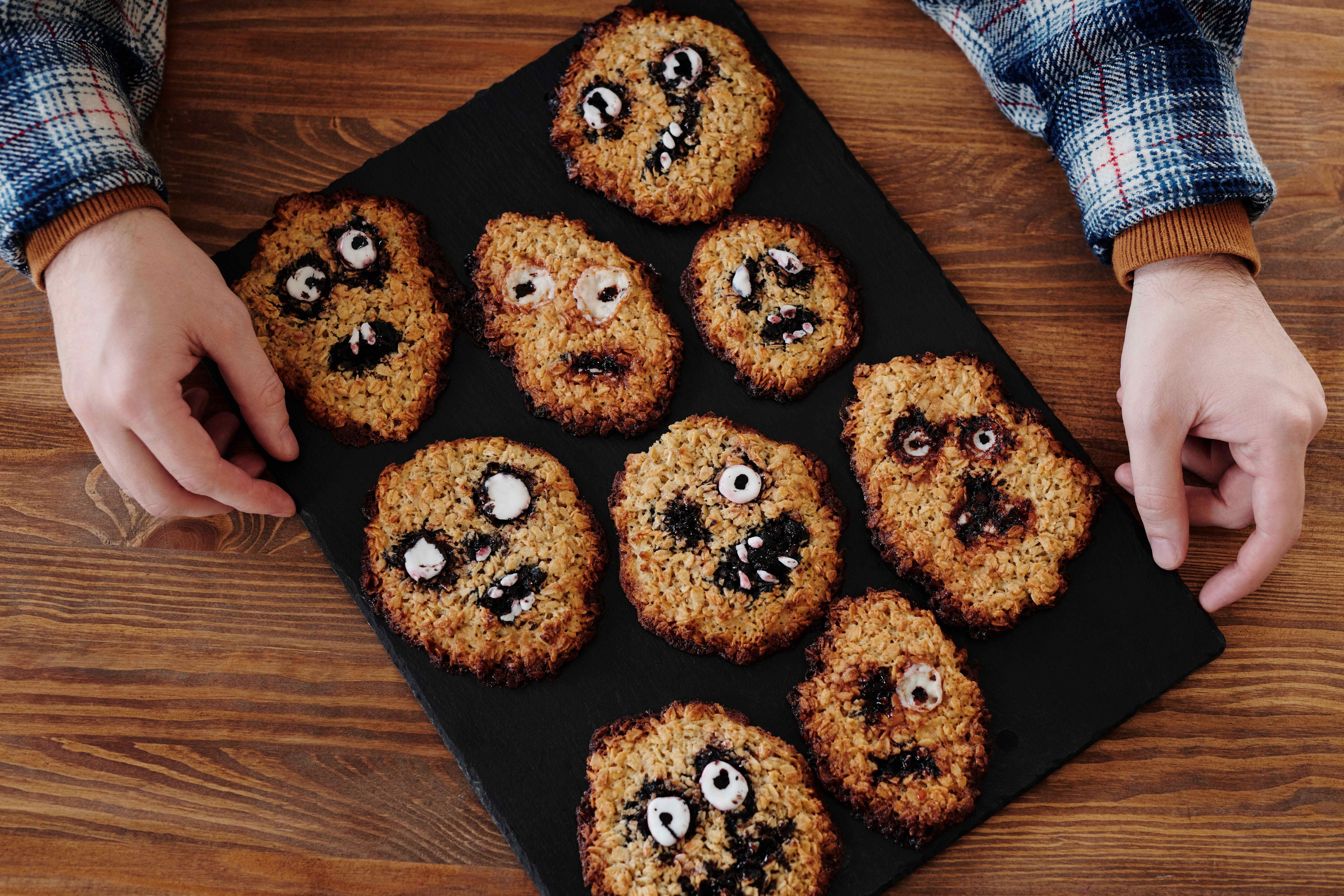 Hands holding a black slate board topped with Halloween oatmeal cookies decorated with dark icing and candy eyes to look like spooky faces.