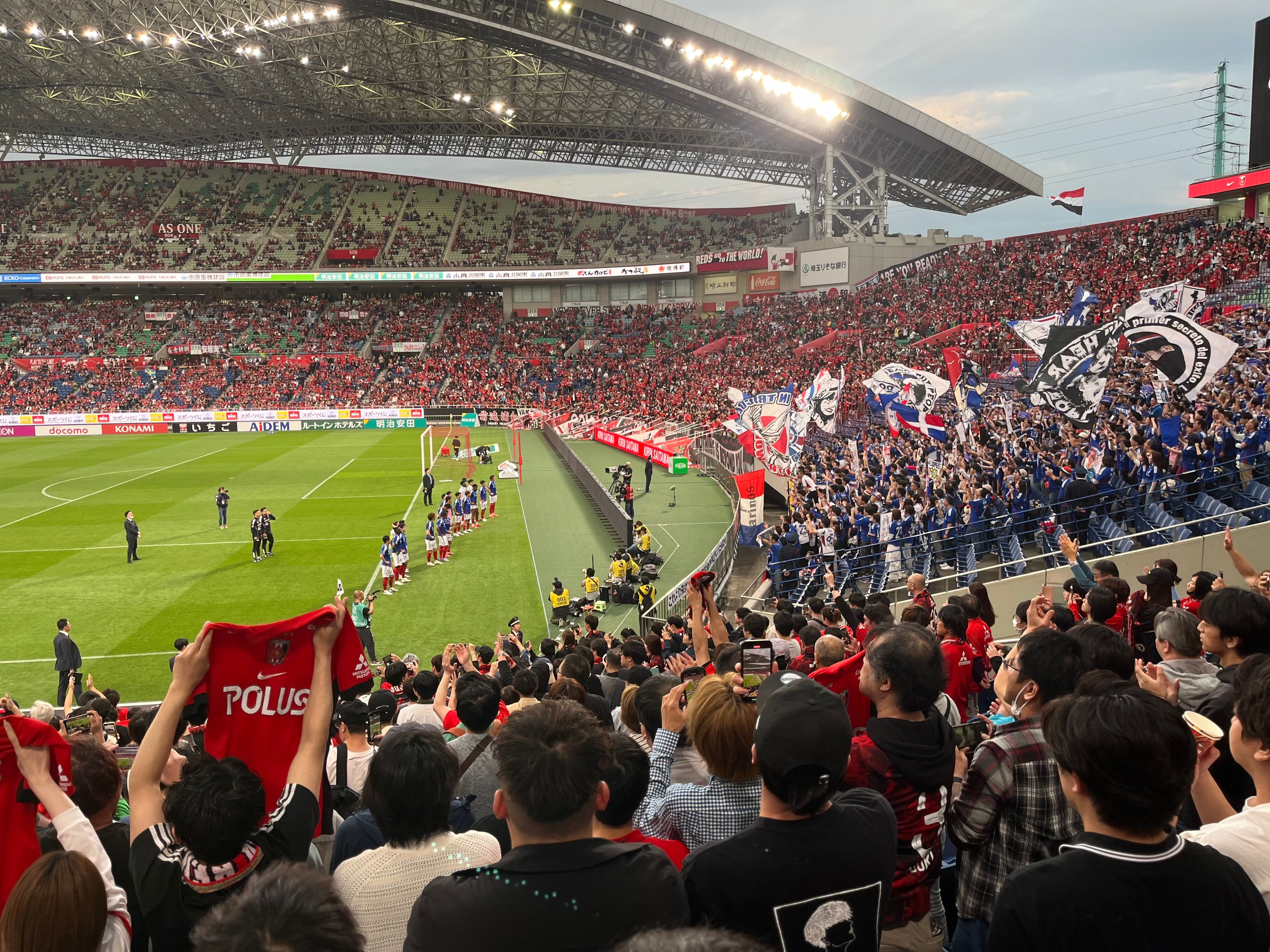 A packed stadium crowd cheering, waving flags, and holding up team jerseys during a soccer match.