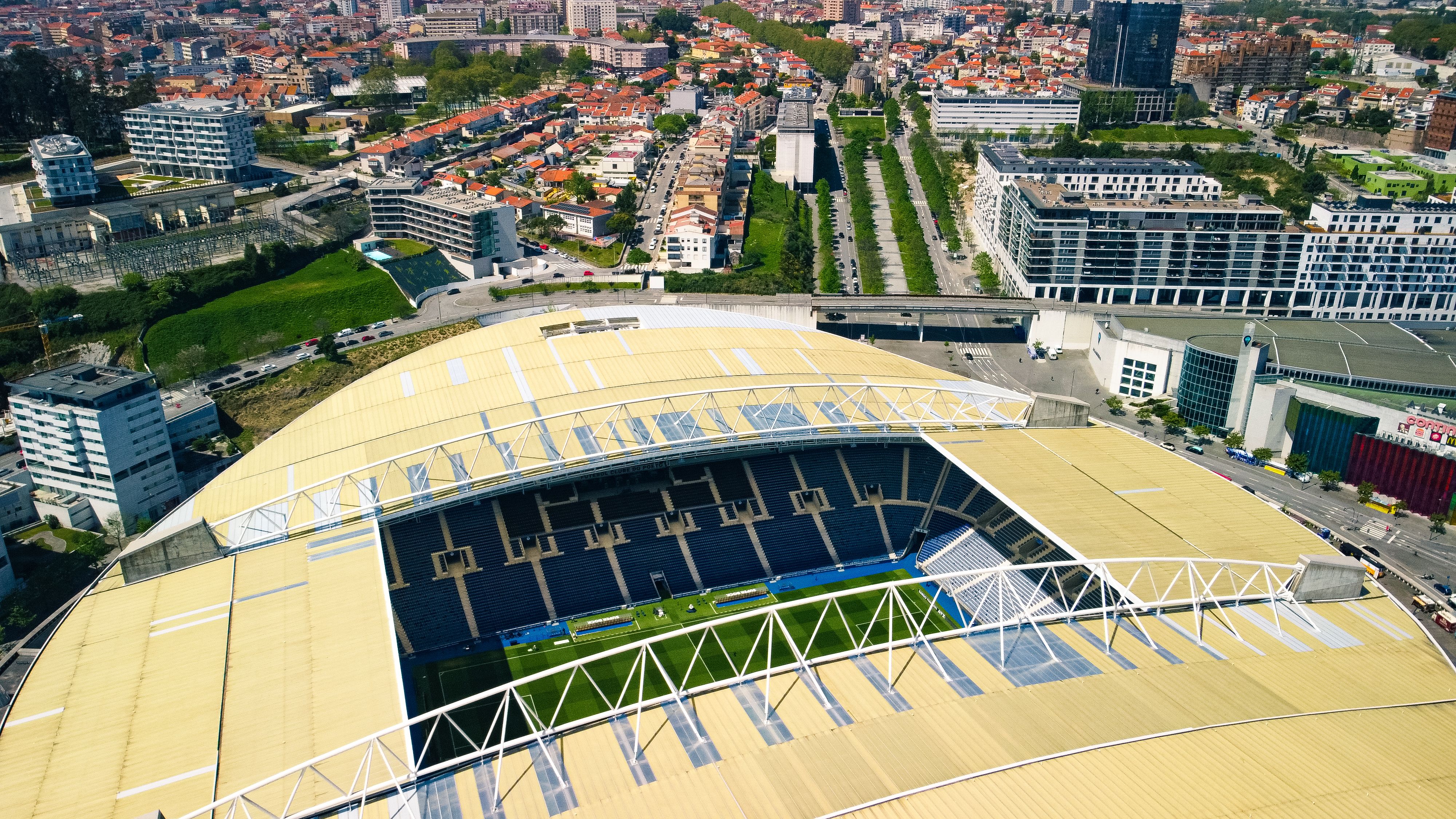 An aerial high-angle view of a modern sports stadium with a yellow wave-like roof structure in an urban setting.