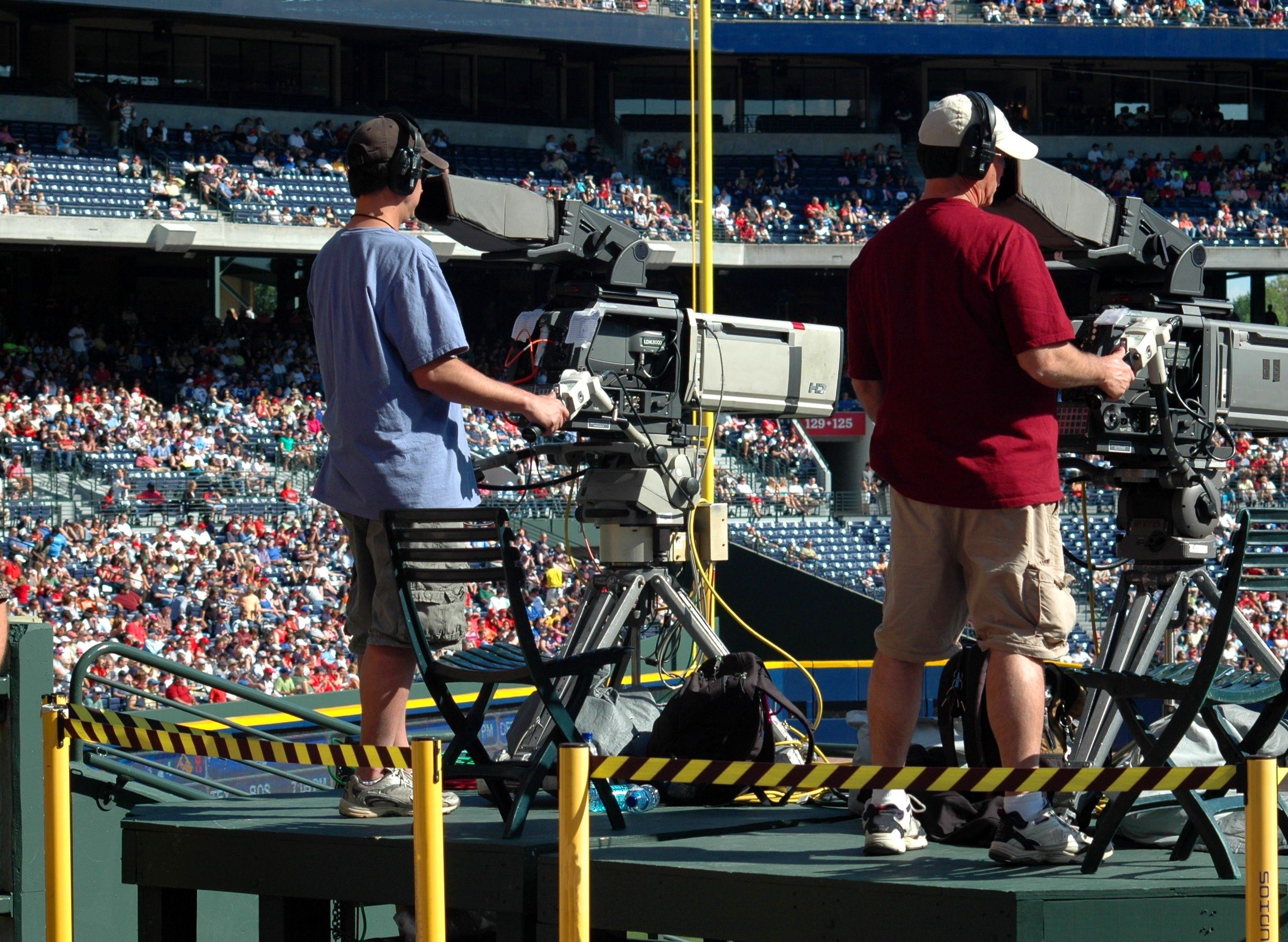 Two camera operators operating large professional broadcast cameras on a raised platform during a stadium sports event.