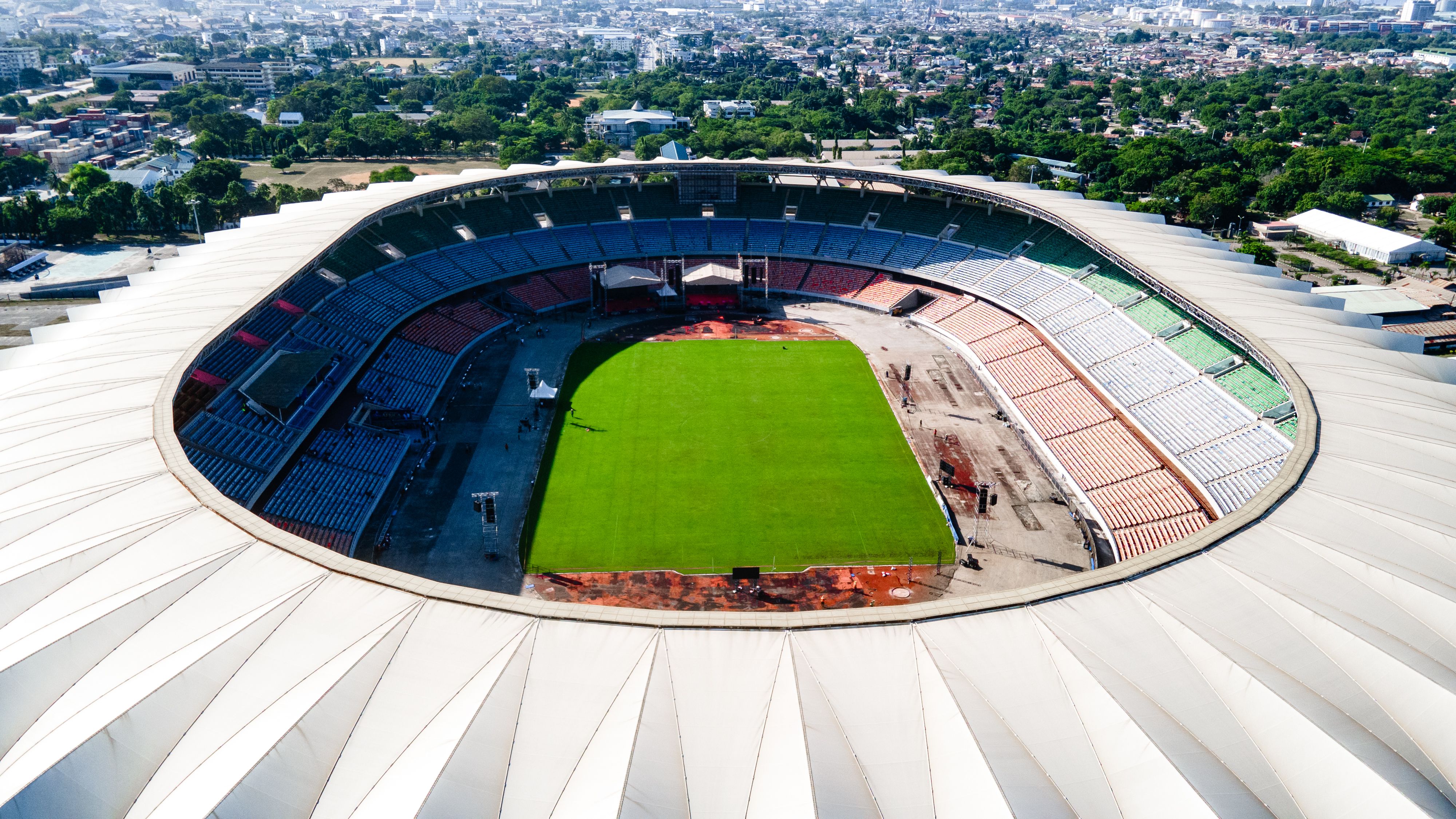 An aerial view of a large sports stadium featuring a green pitch surrounded by colorful seating tiers and urban scenery.