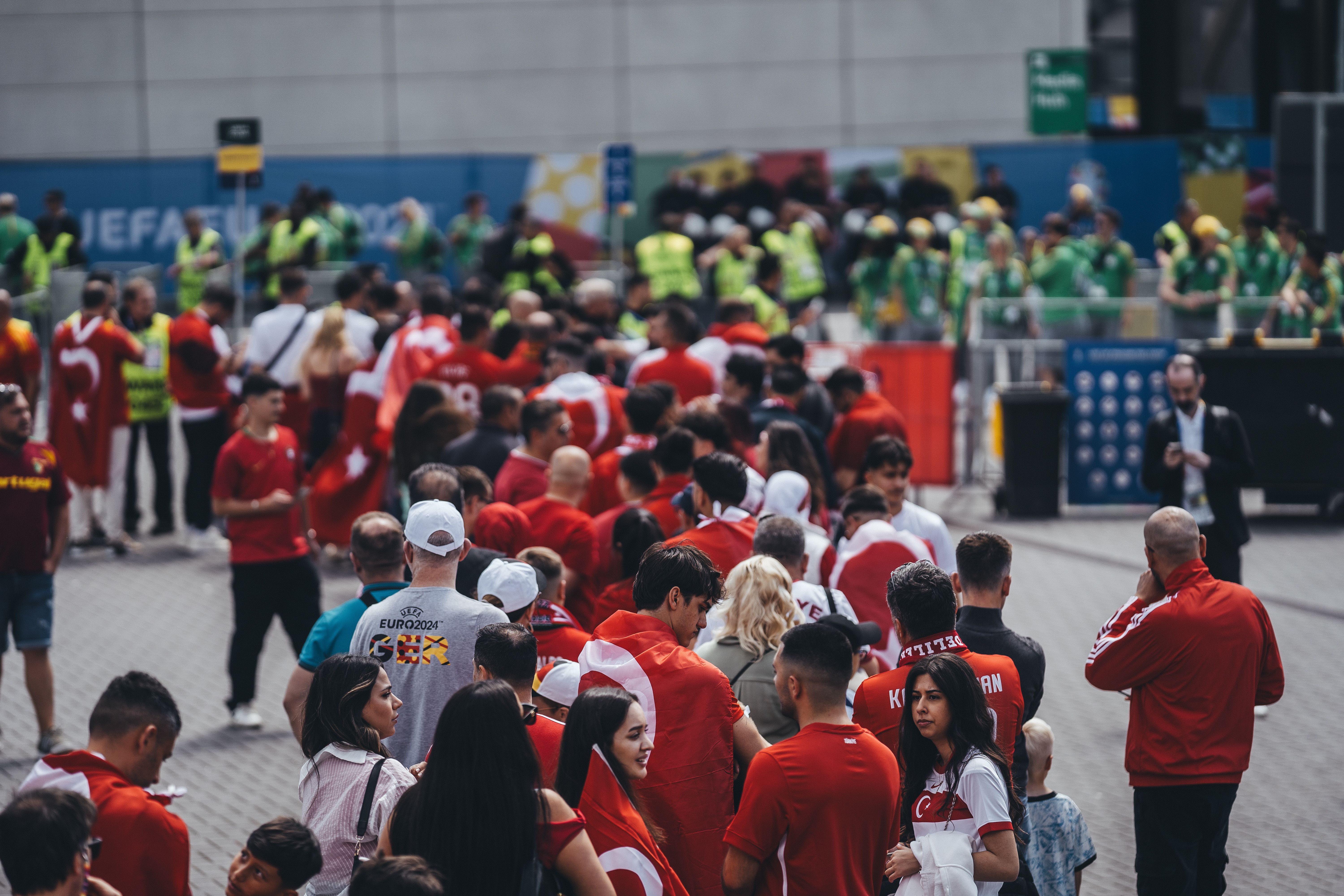 A large crowd of soccer fans wearing red shirts and draped in national flags walking toward an event entrance venue.