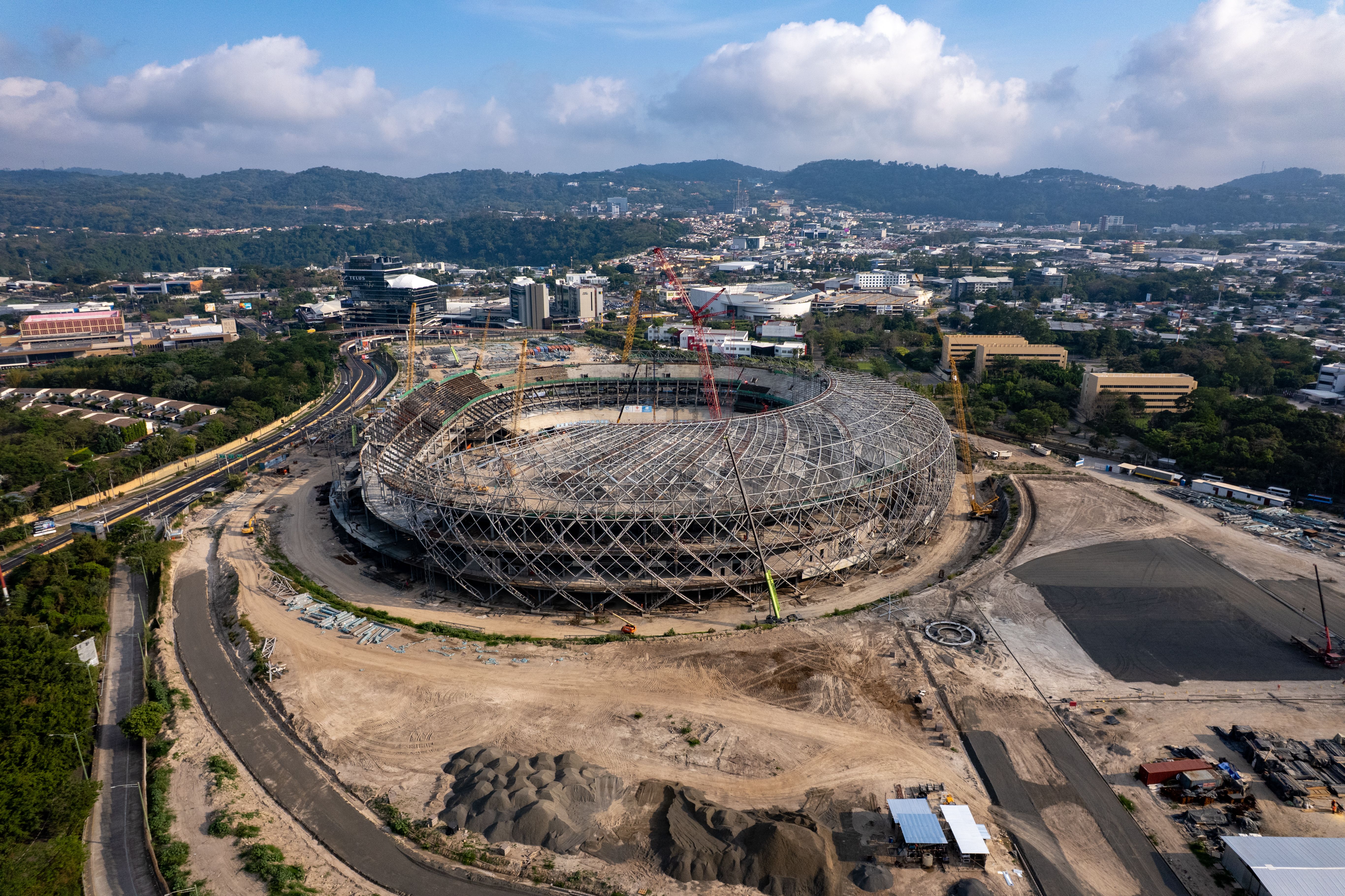 Aerial view of a massive stadium under construction with heavy cranes, steel framework, and surrounding city landscape.