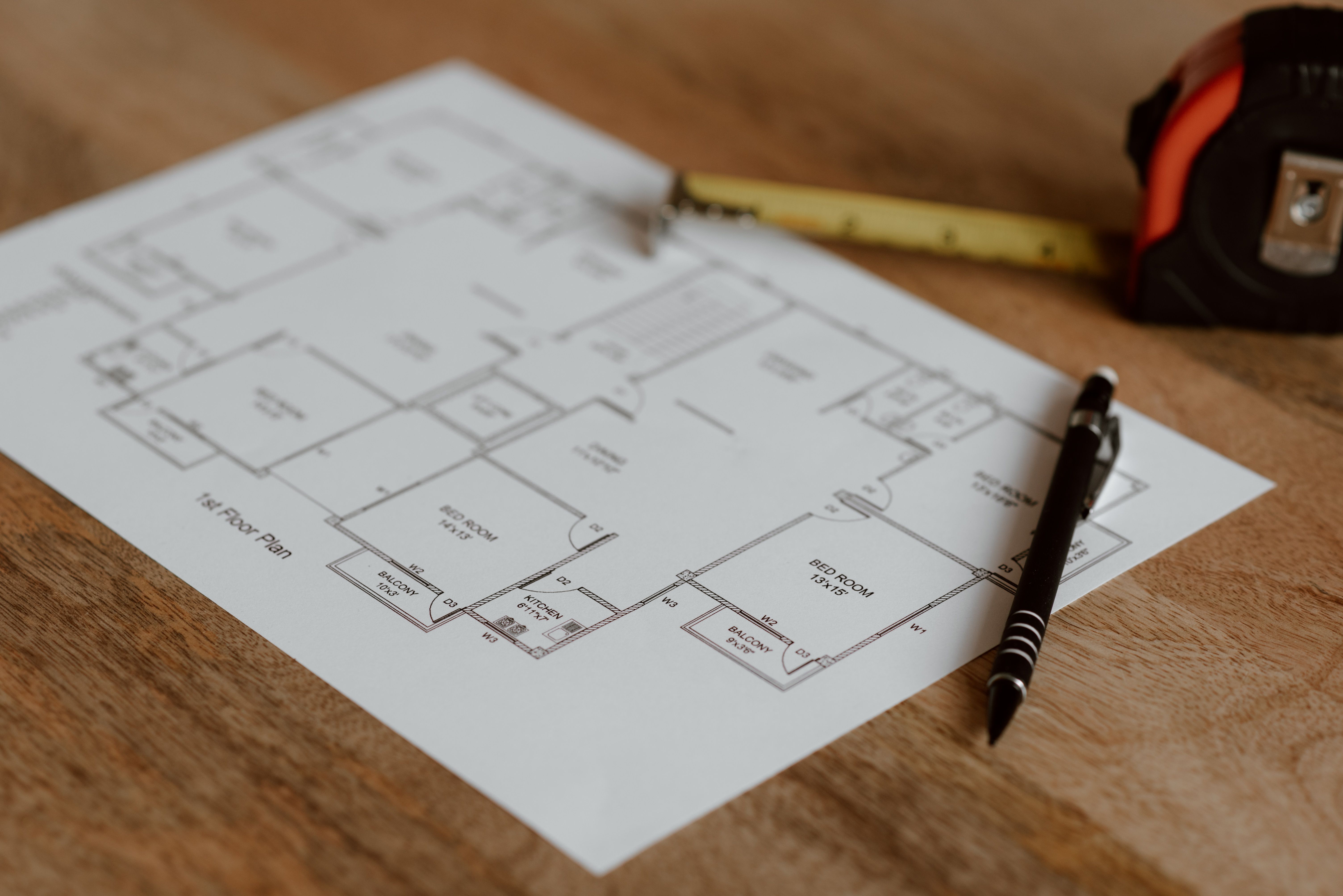 A printed architectural first-floor house plan resting on a wooden surface with a pen and a red tape measure alongside it.