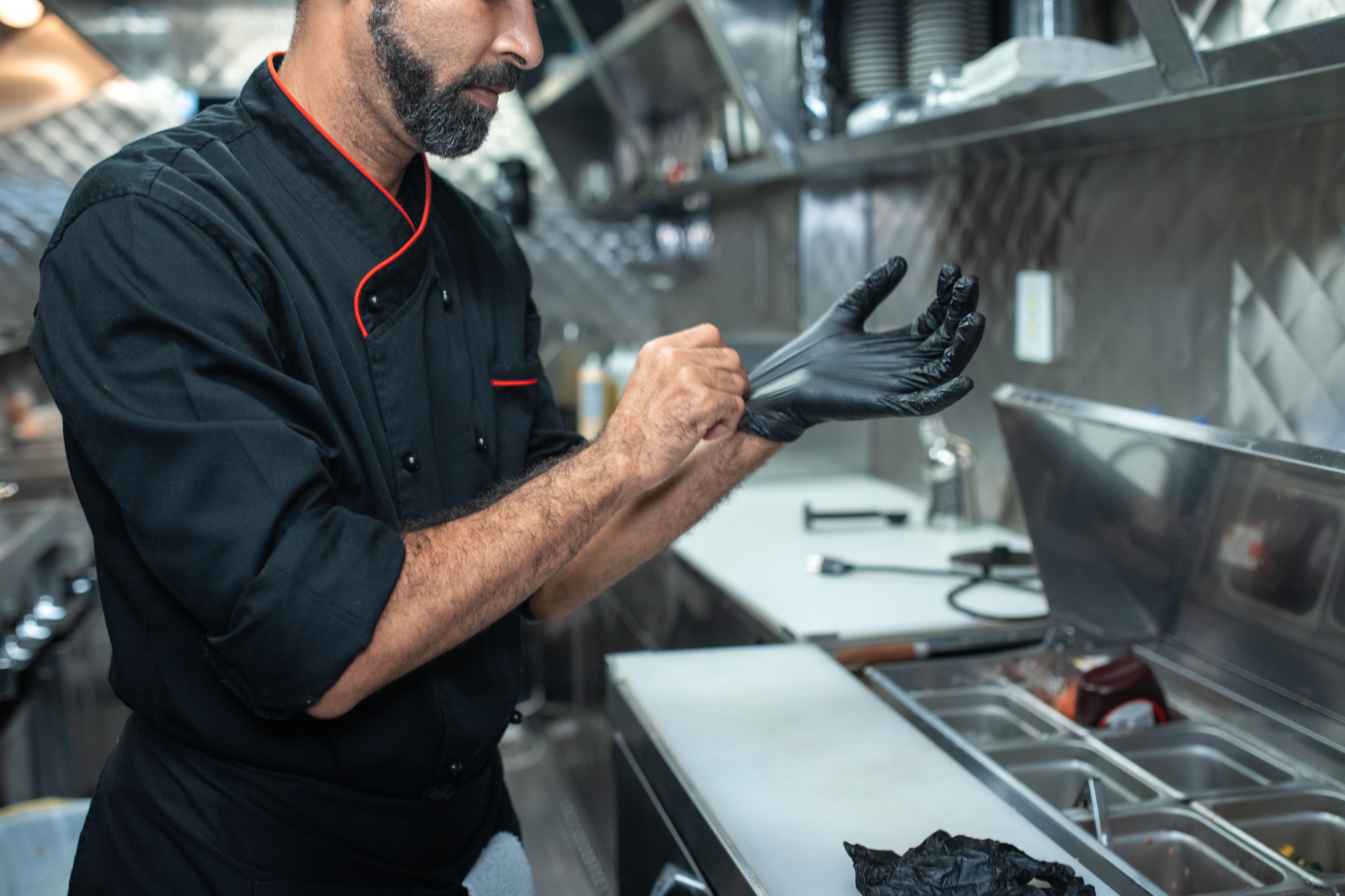 A male chef in a black uniform putting on protective black disposable gloves in a commercial kitchen prep area.