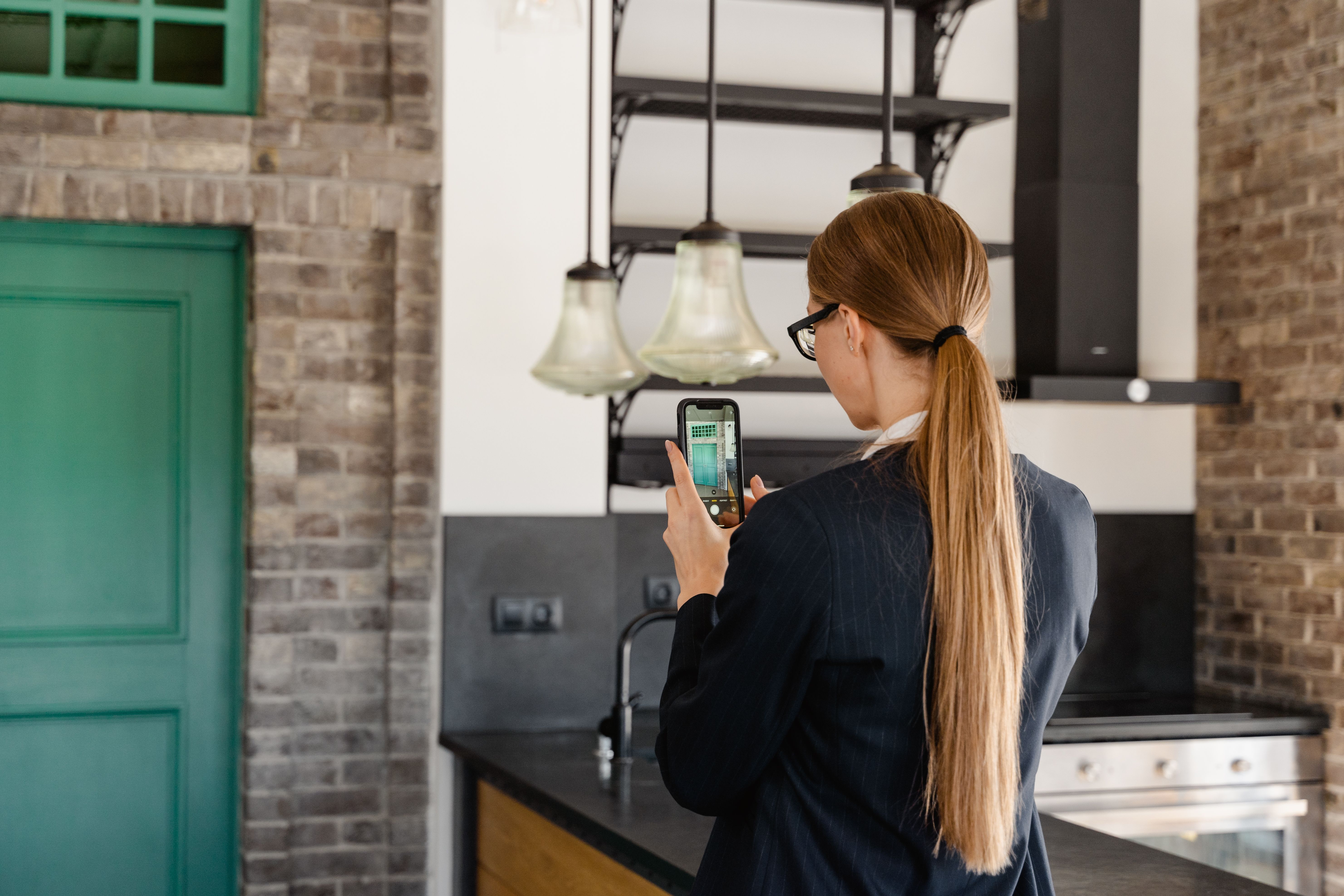 A woman with long hair taking a photo of a kitchen interior on her smartphone during a facility inspection.
