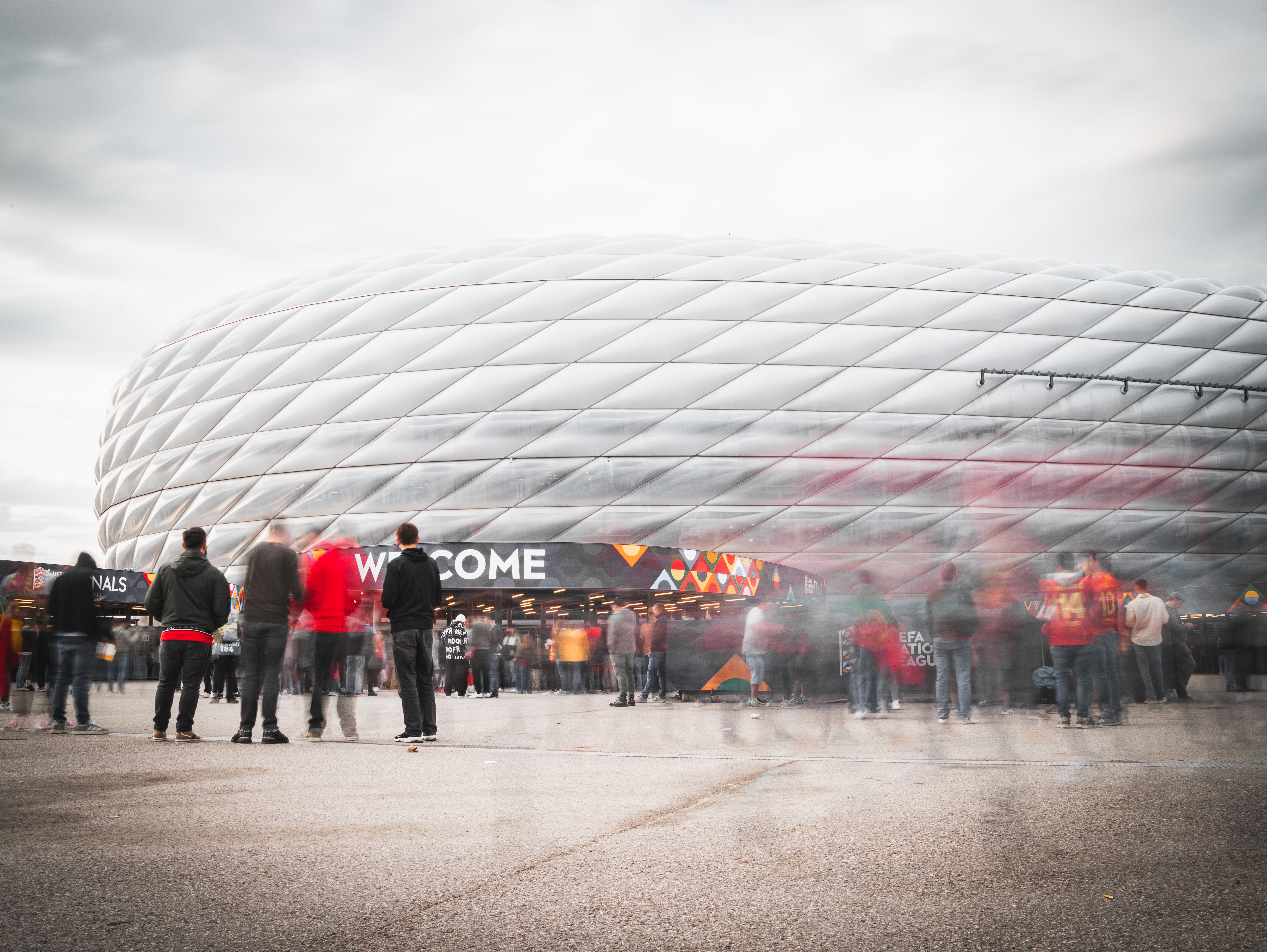 Crowds of sports fans gathering near the entrance gate outside a large modern stadium on an overcast day.
