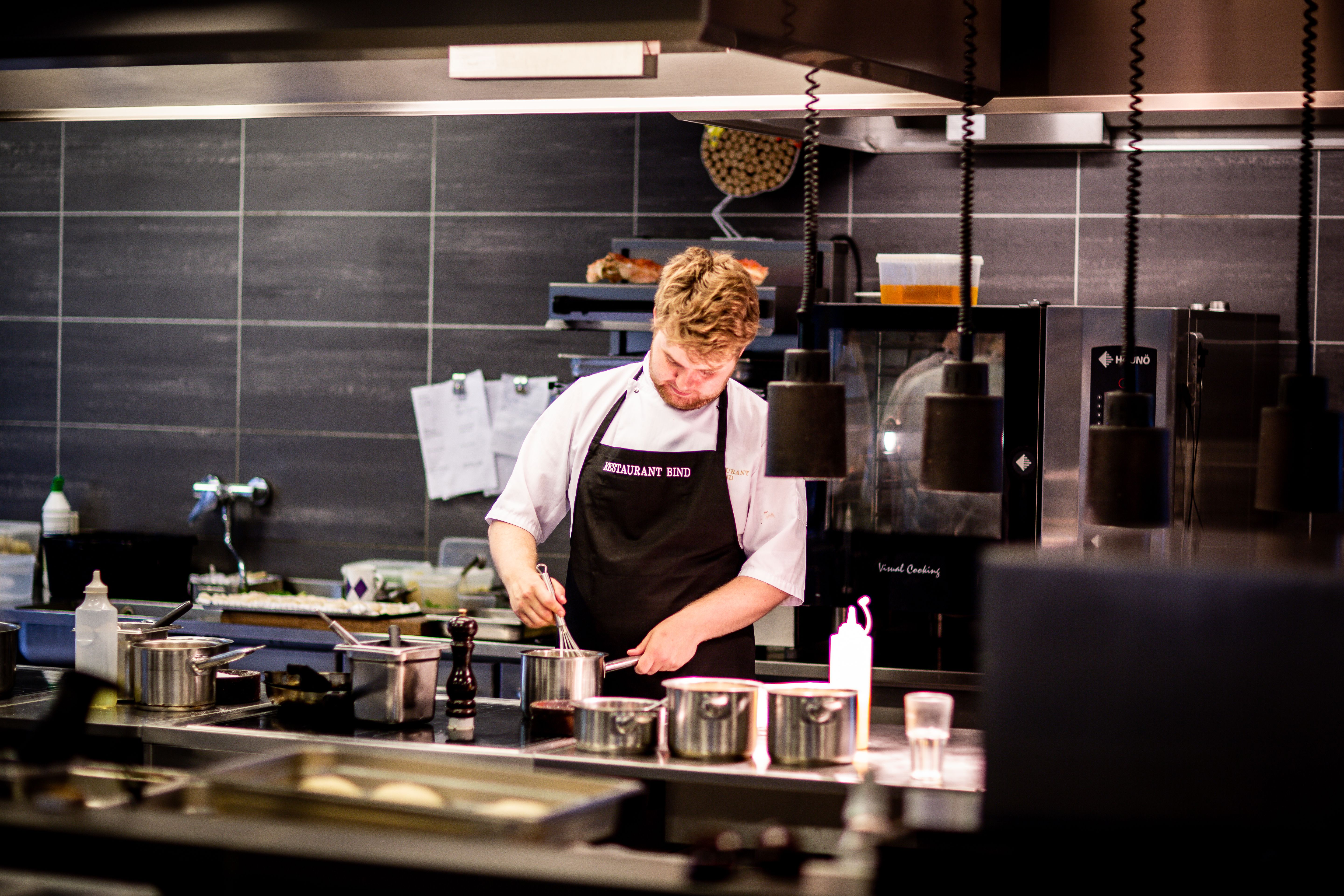 A male chef in a black apron whisking ingredients in a stainless steel pot inside a commercial restaurant kitchen.