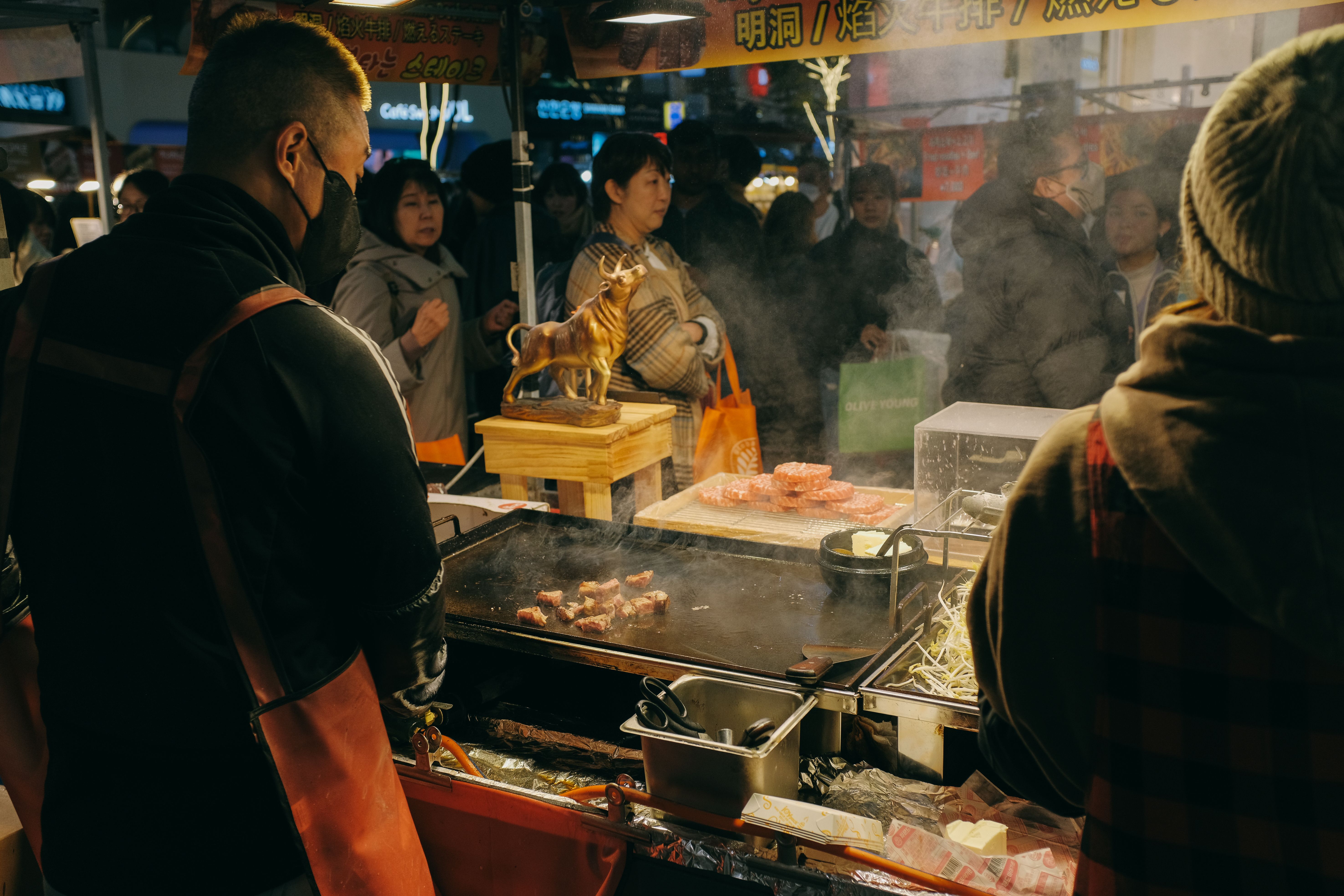 A street food vendor grilling meat on an outdoor flat-top grill at a busy night market counter.