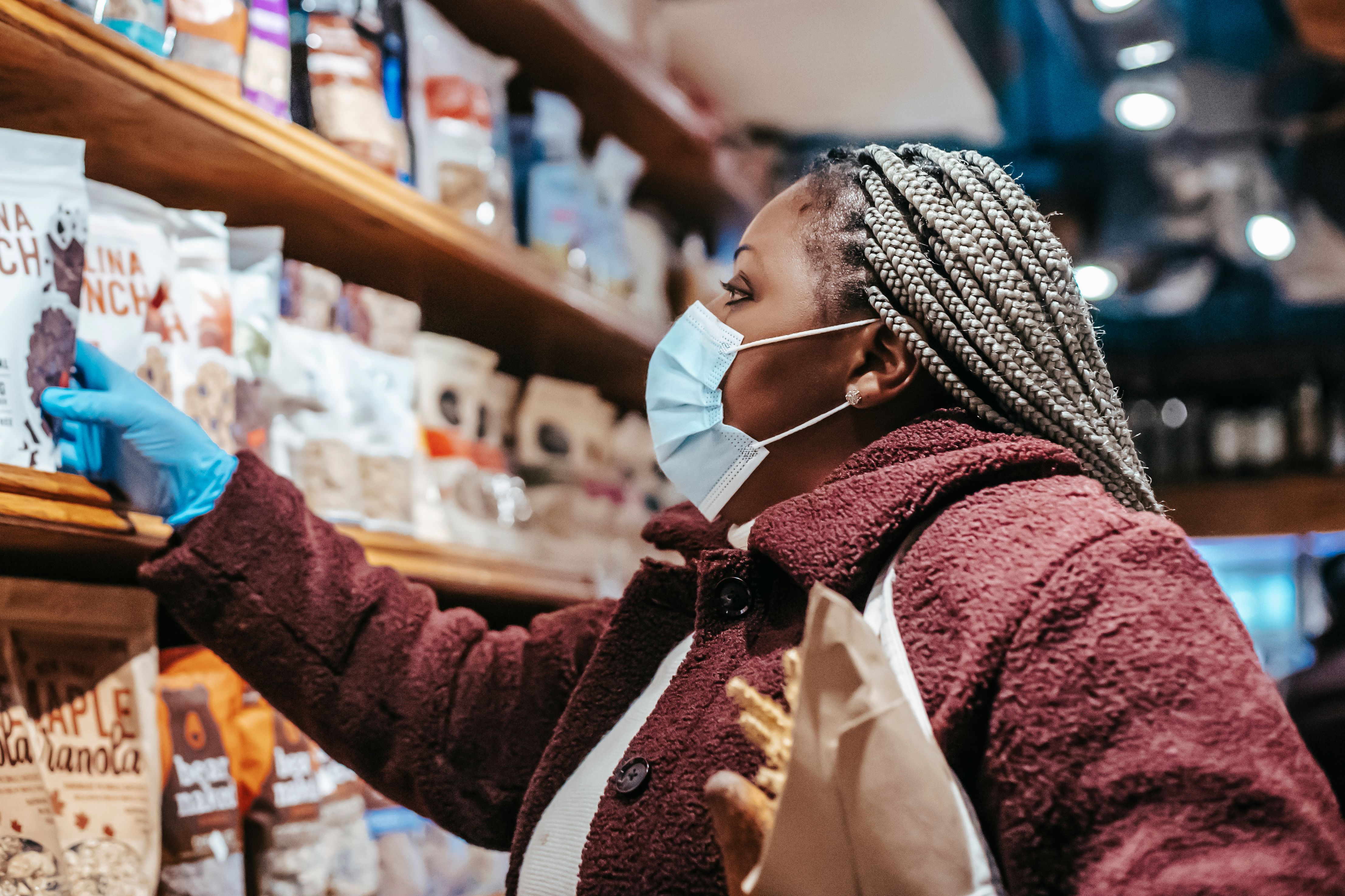 Woman wearing a face mask and protective gloves browsing packaged snacks on wooden grocery store shelves.