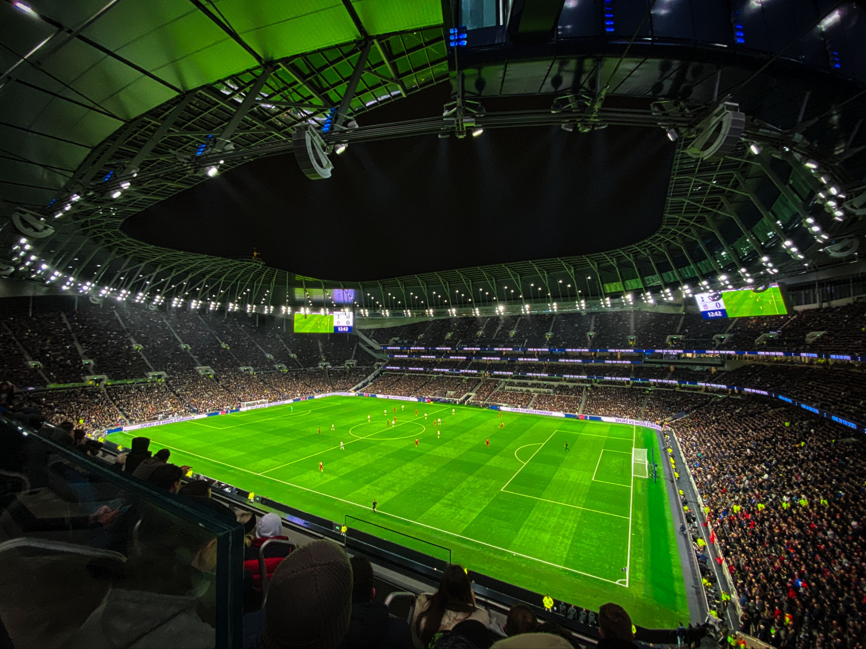 Wide interior view of a packed modern football stadium lit up at night with a bright green pitch and spectators.