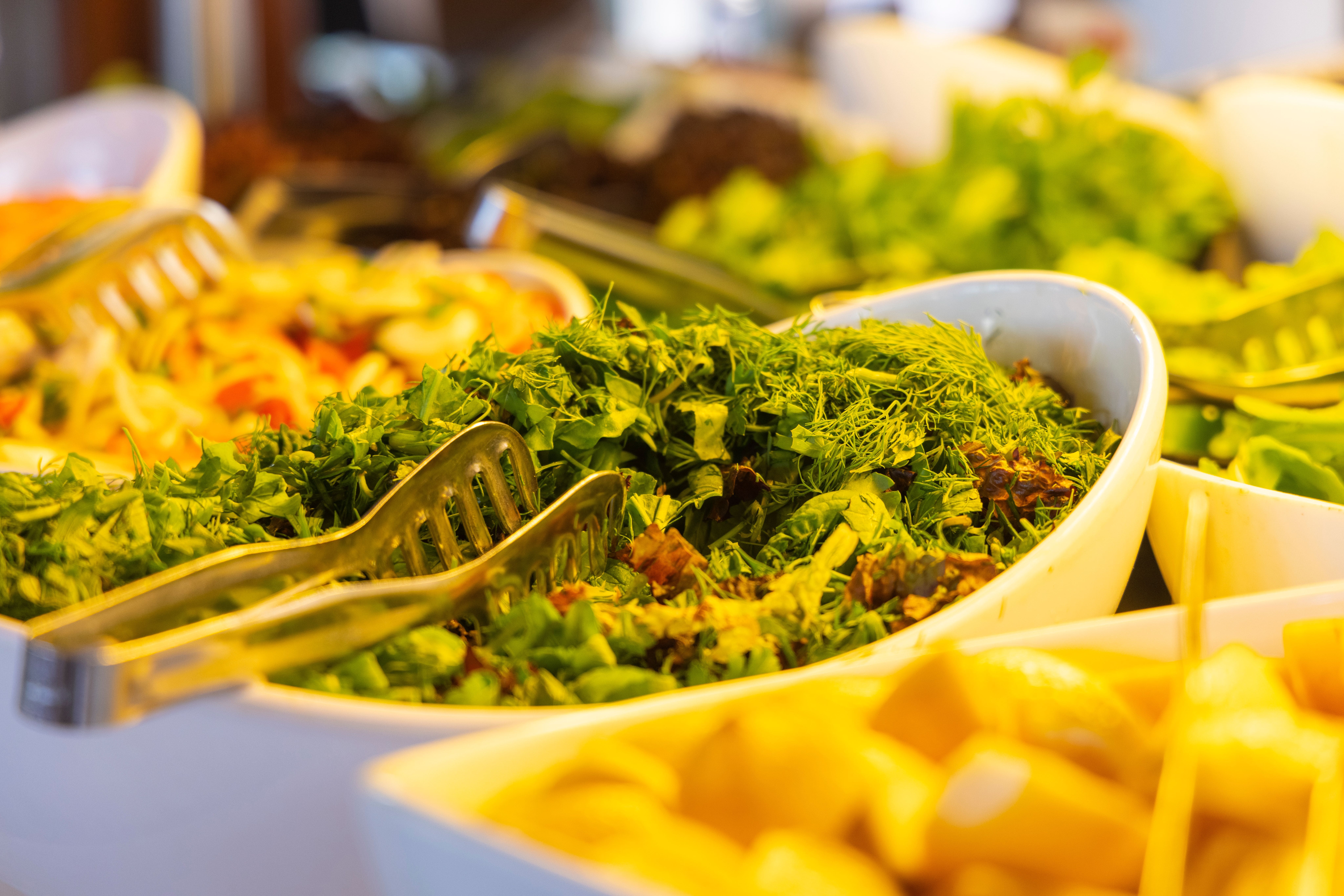 Close-up of fresh green chopped herbs in a white serving dish with metal tongs on a buffet spread.