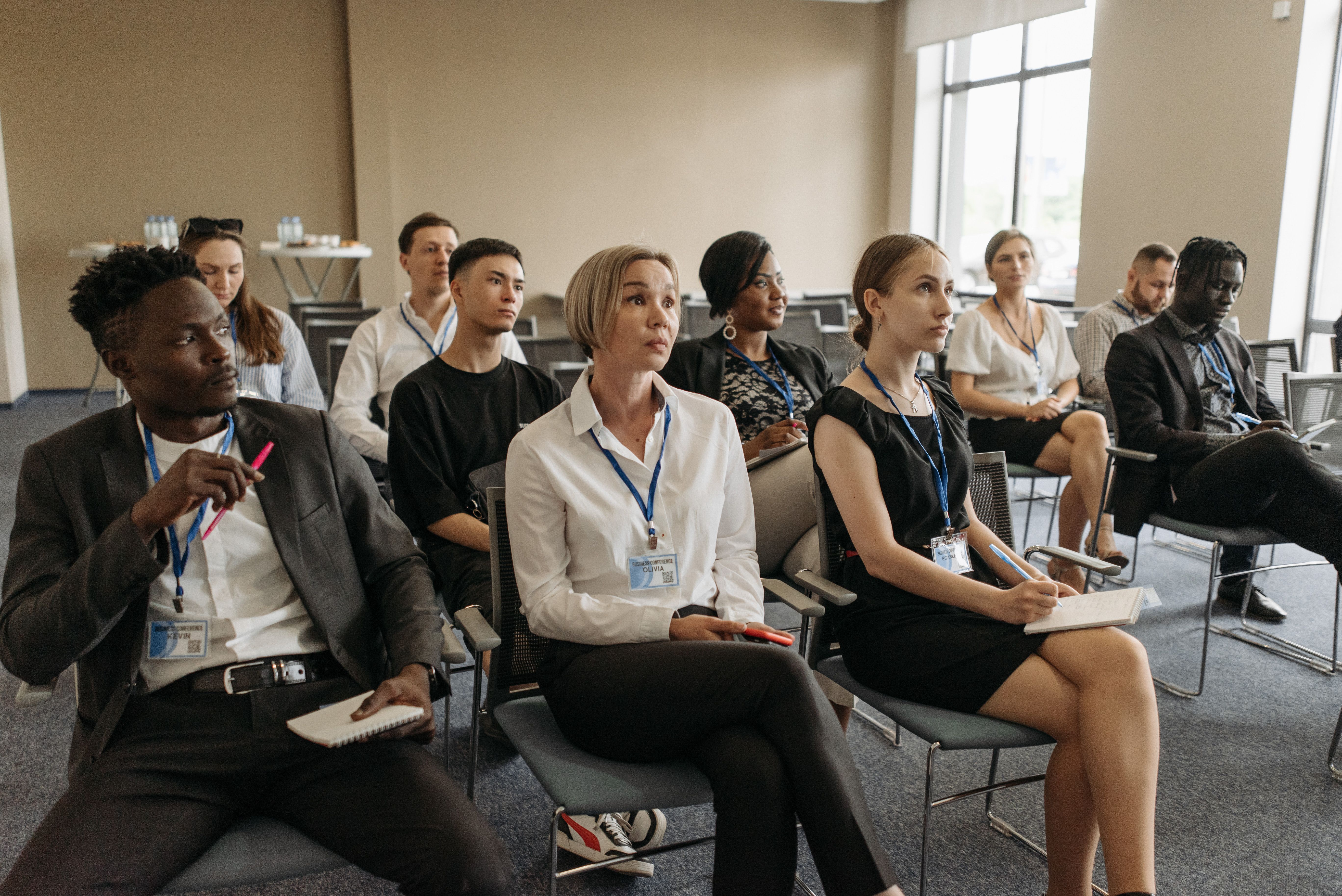 Business professionals sitting in rows of chairs at a seminar while taking notes during a presentation.