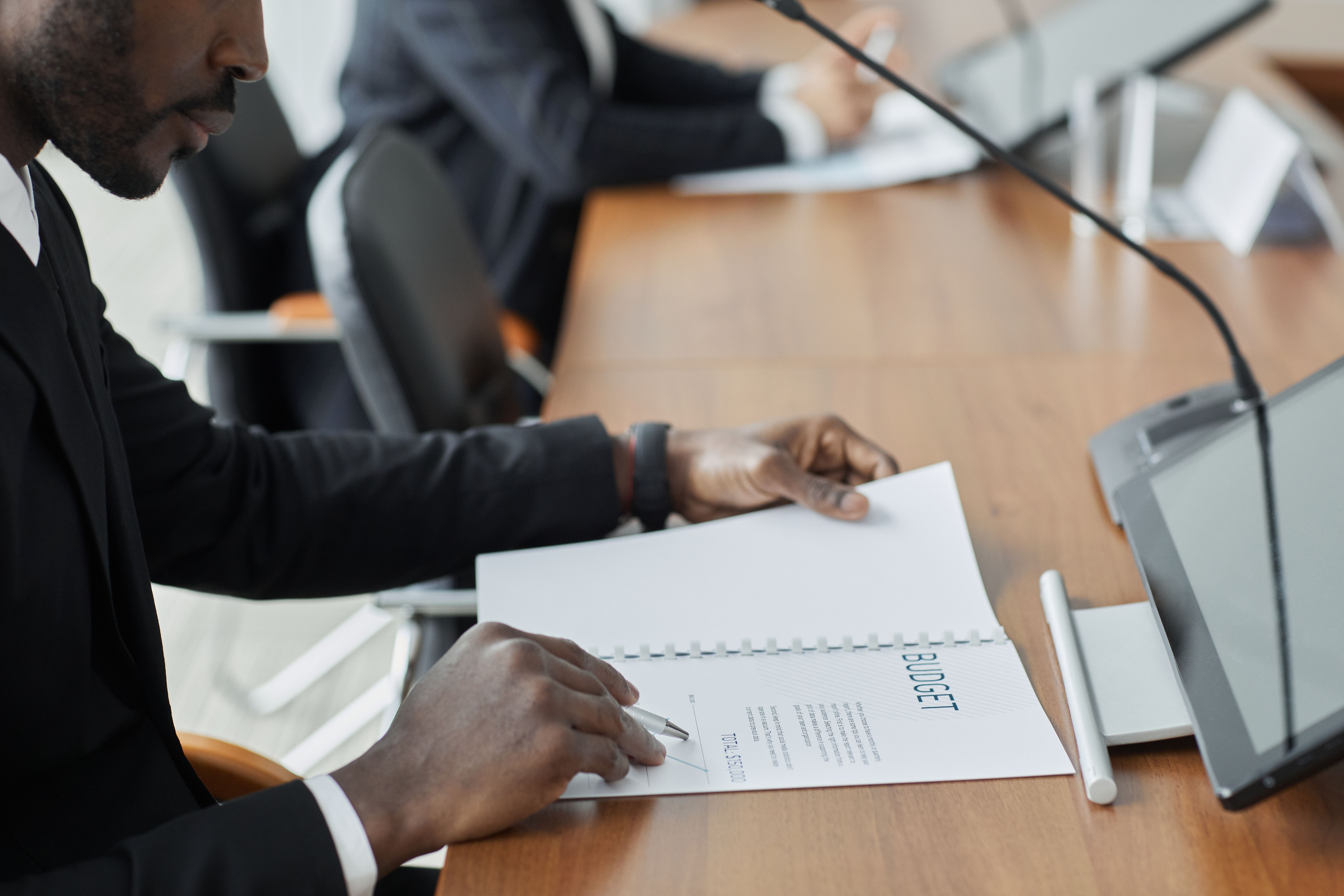 A businessman sitting at a conference table reviewing and signing a printed budget document with a pen.