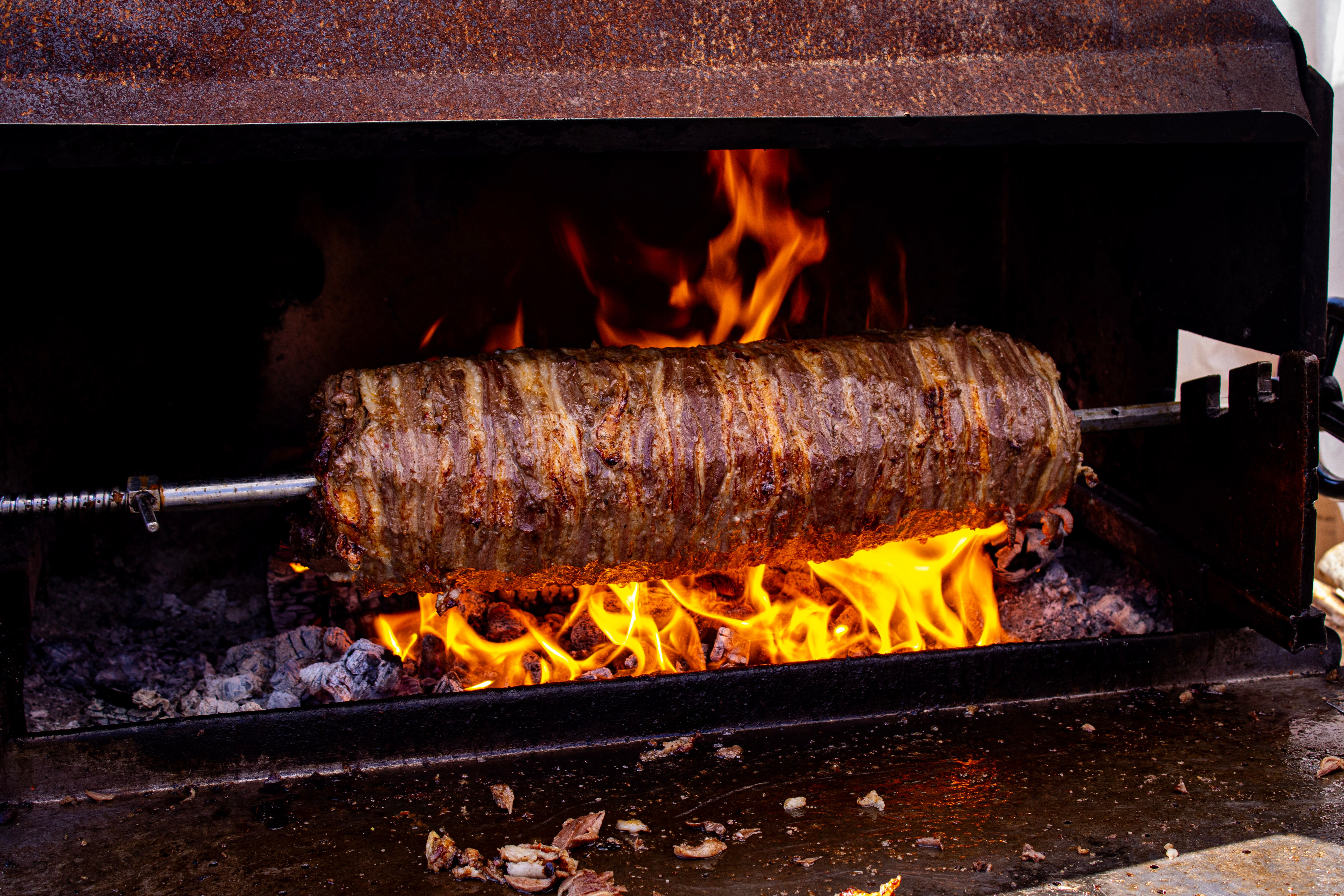 Large roll of seasoned doner kebab meat rotating on a horizontal spit over open flames in a commercial rotisserie grill.