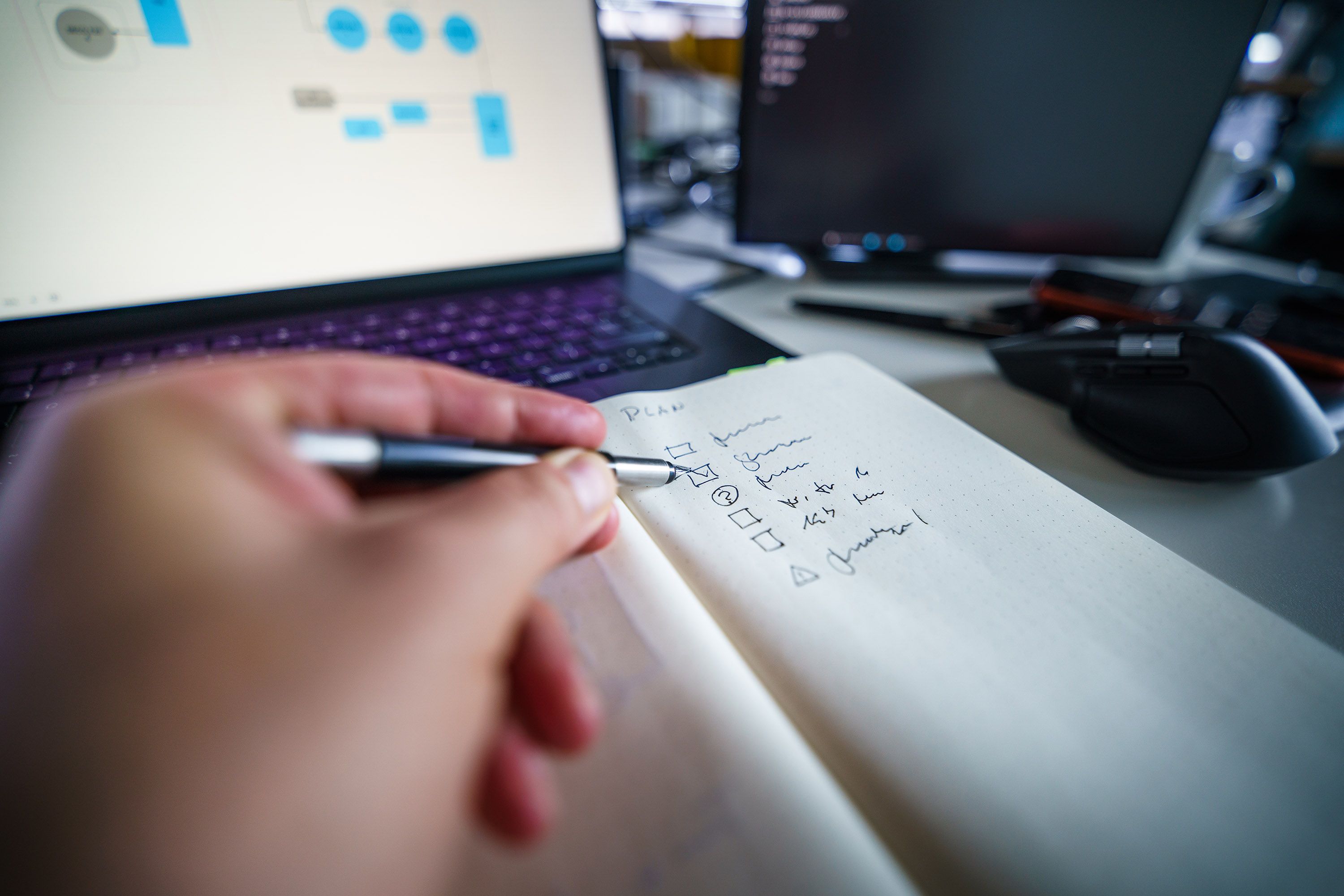Close-up of a person marking off checkboxes in a handwritten notebook planner next to a laptop displaying workflow diagrams.