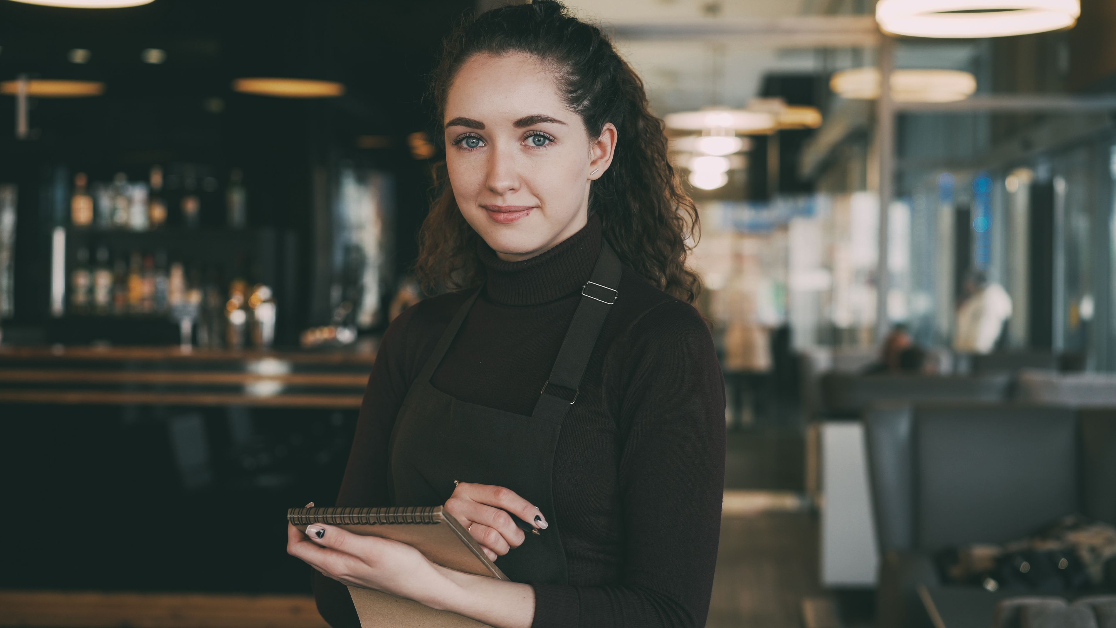 A waitress in a brown apron holding a notepad and pen while smiling in a restaurant setting.