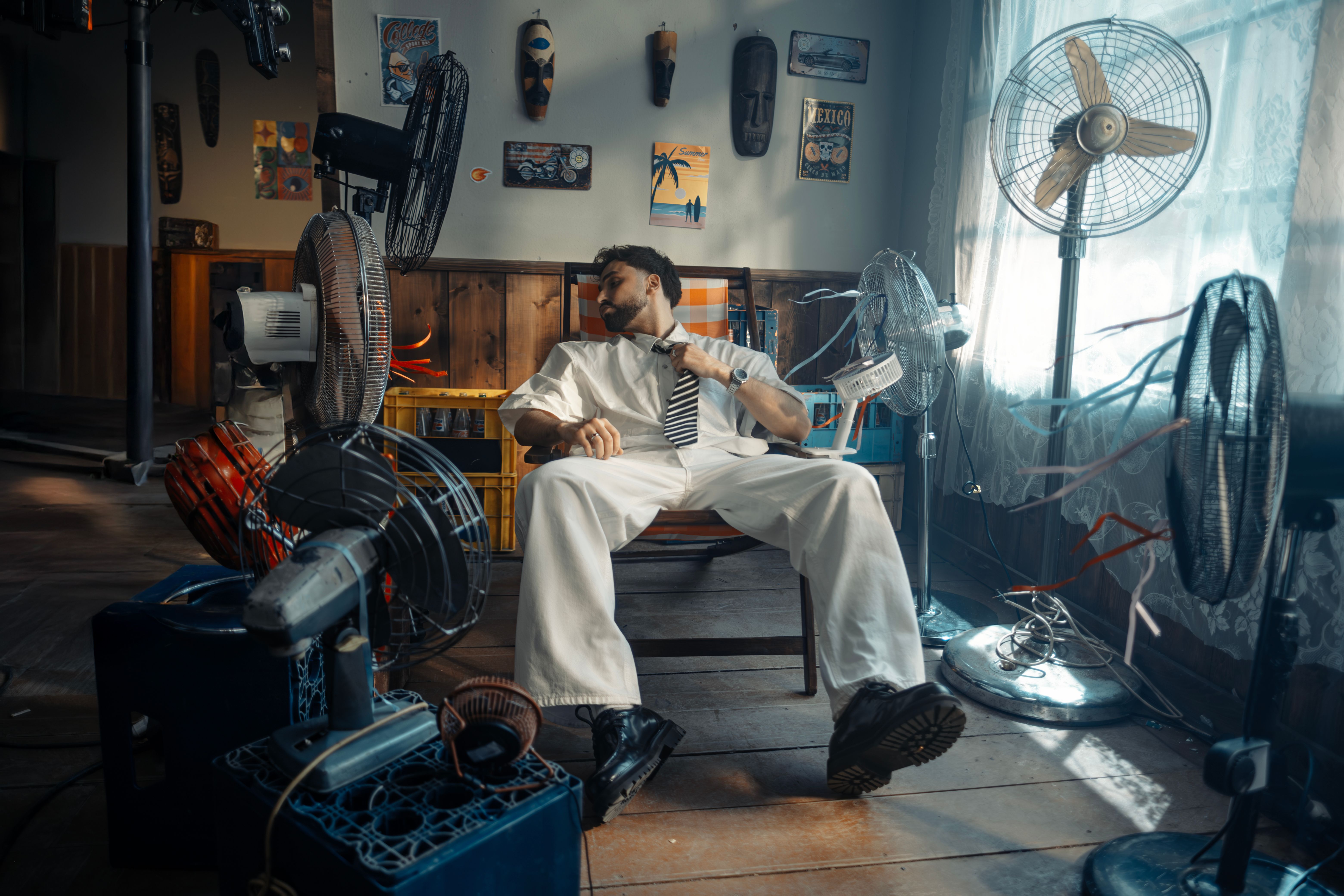 A man loosening his tie and resting in a chair while surrounded by multiple electric fans blowing air on him.