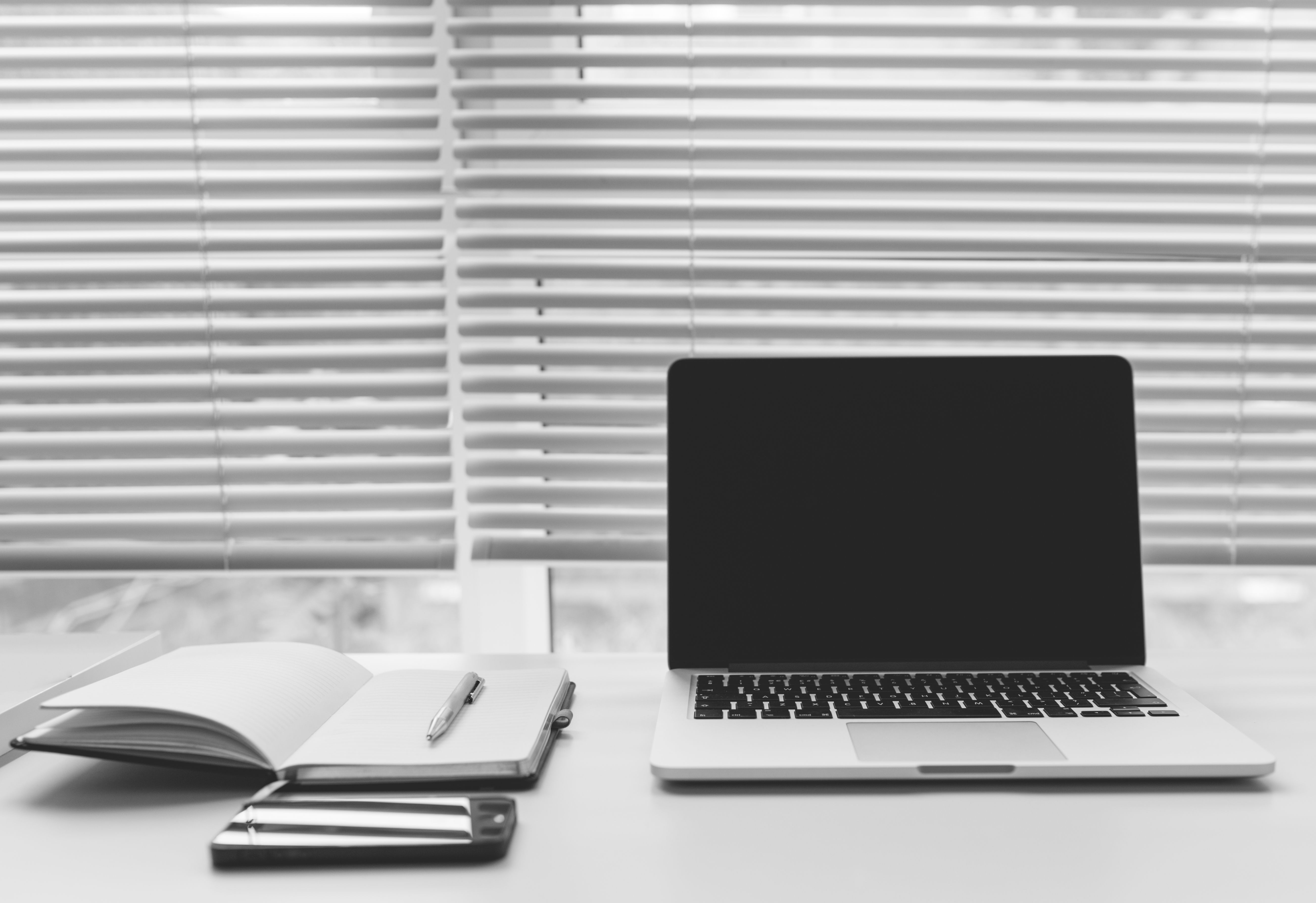 Black and white view of a minimalist desk setup with an open laptop, notepad, pen, and smartphone in front of window blinds.