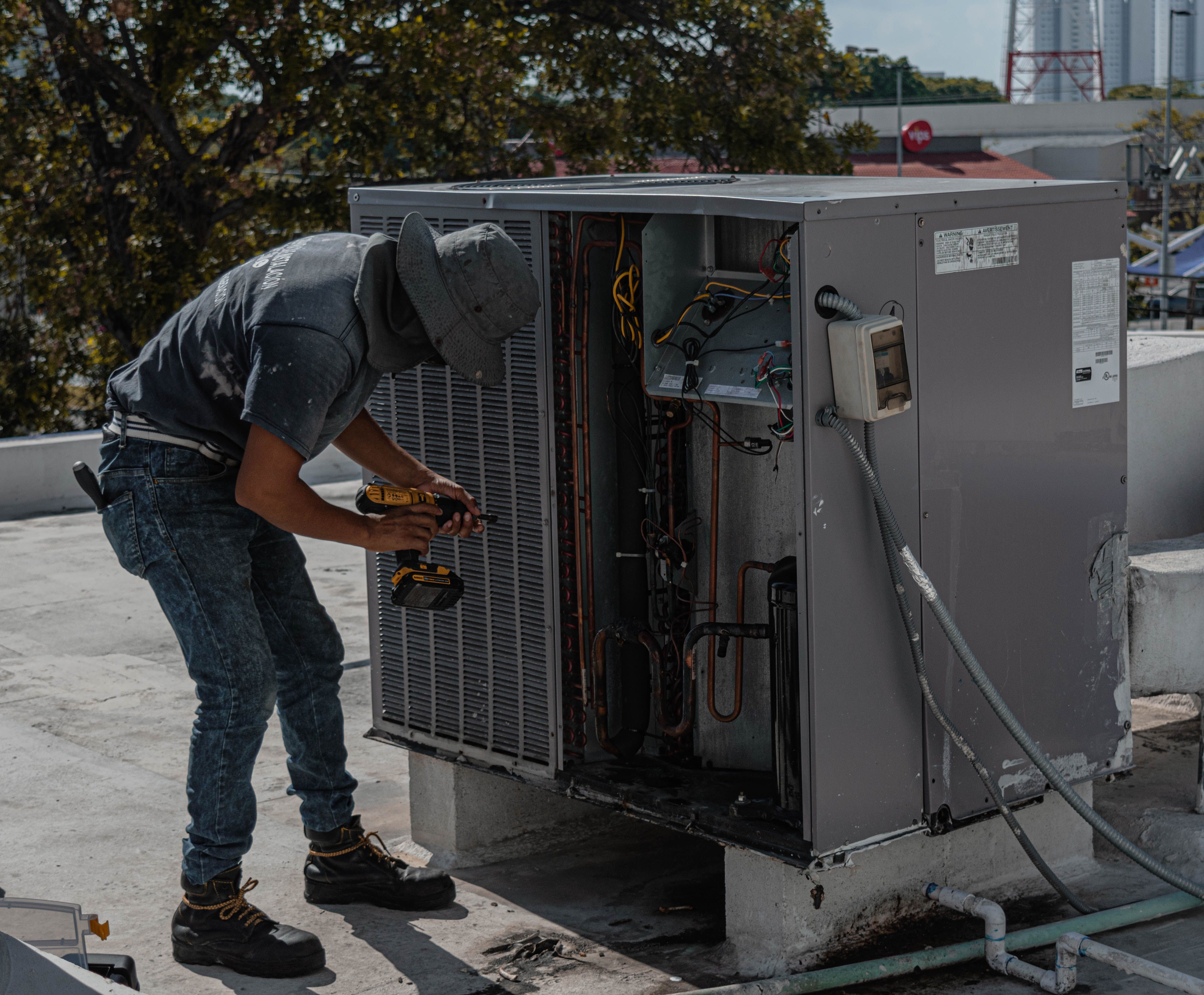 An HVAC technician using a power drill to repair an outdoor air conditioning unit on a rooftop.