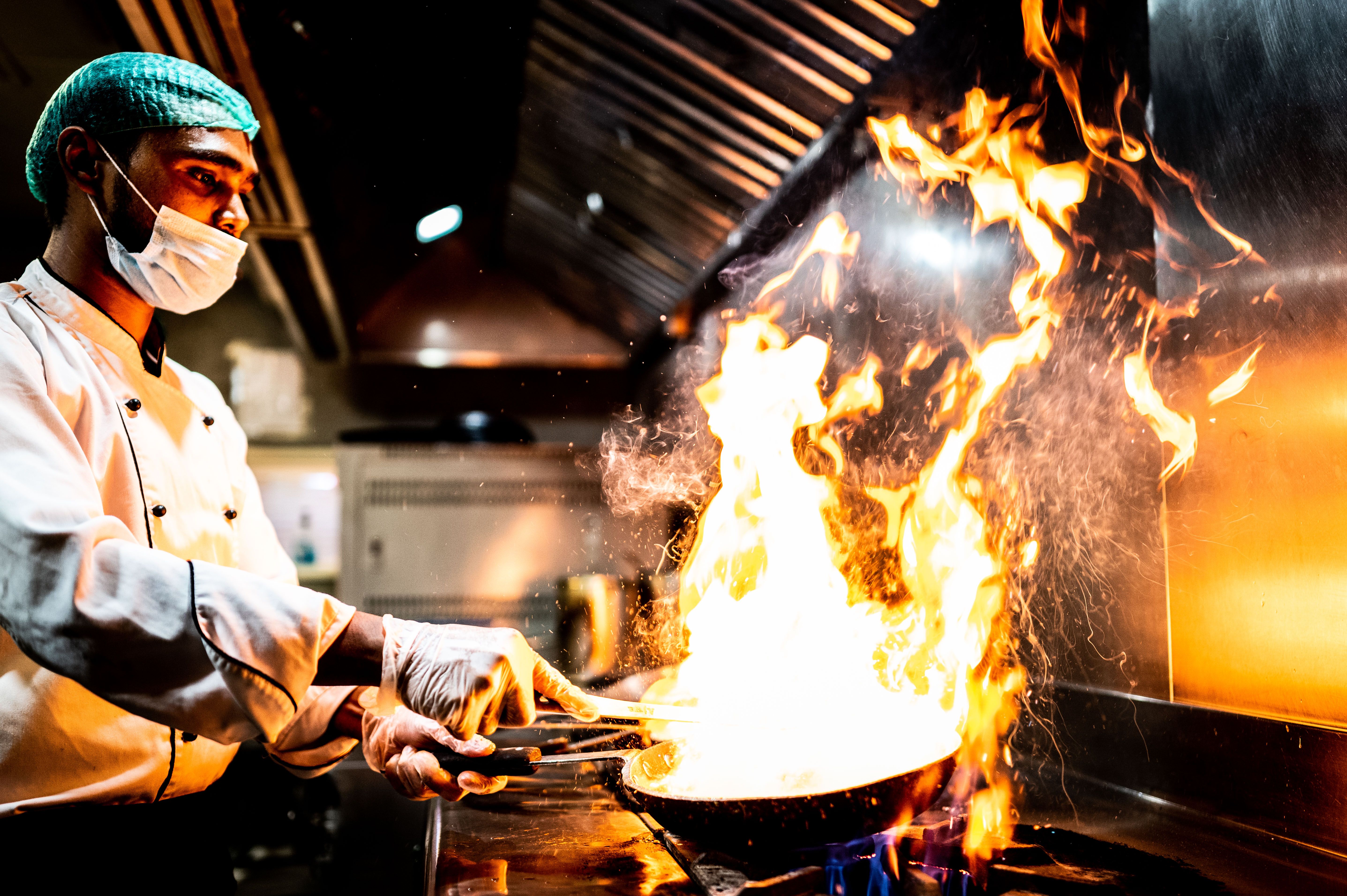 A professional chef in a hairnet, face mask, and gloves cooking with high flames in a pan at a commercial kitchen stove.