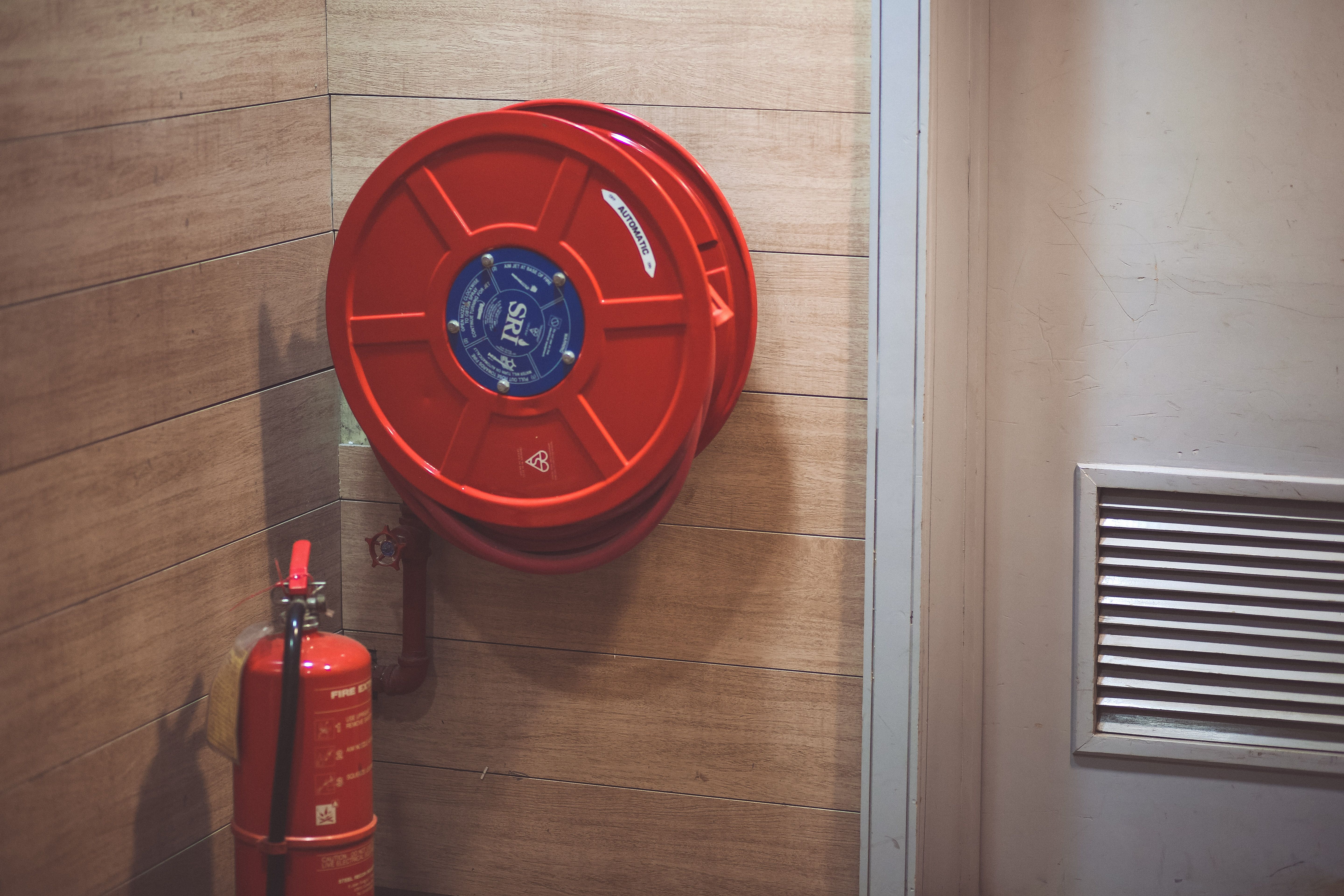 A red wall-mounted fire hose reel and portable fire extinguisher installed against a wooden panel wall next to a ventilation grille.