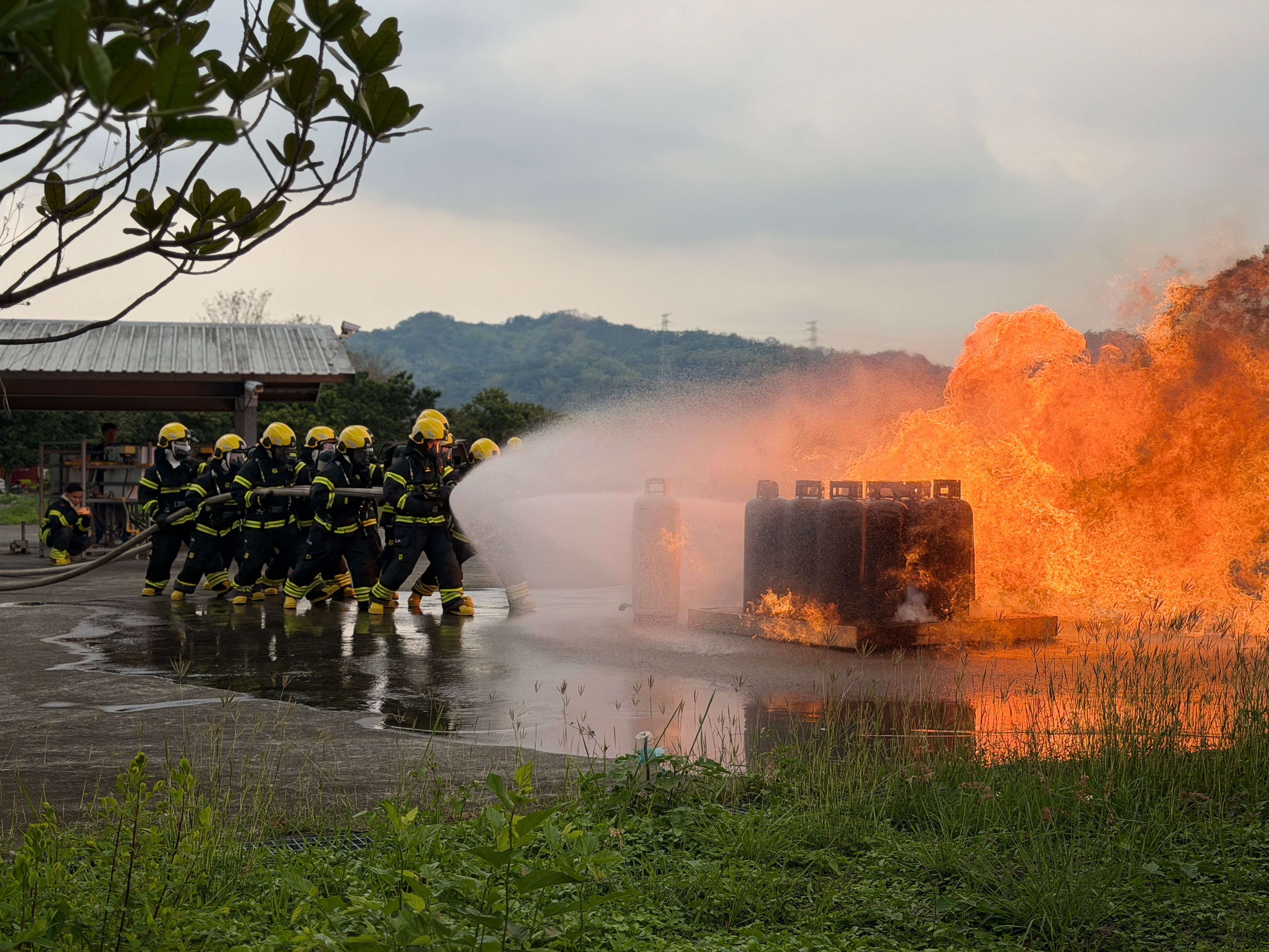 A team of firefighters in protective gear spraying water from a hose to extinguish a large fire during a outdoor safety training exercise.