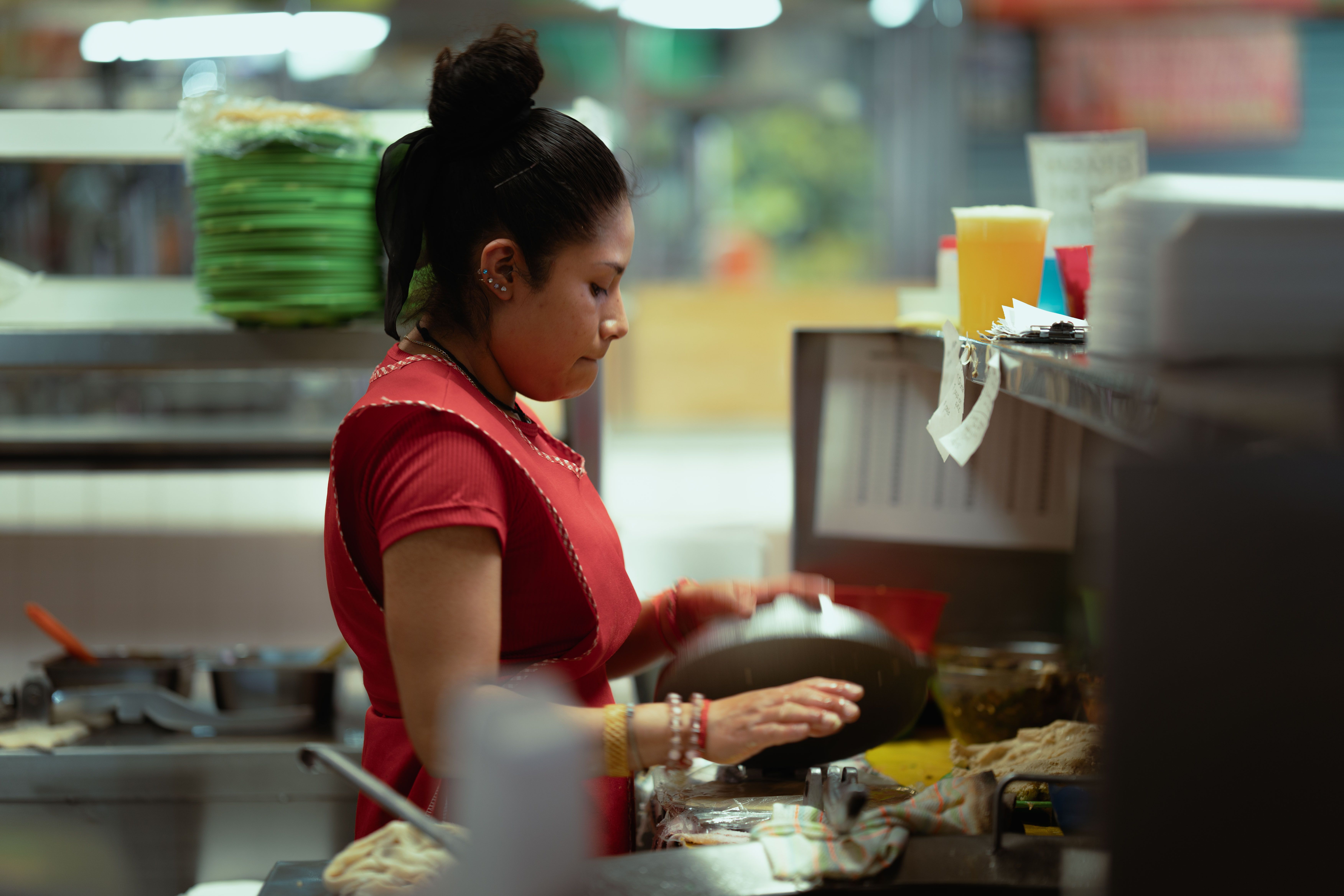 Side view of a female kitchen staff member in a red apron preparing food behind a commercial food service counter.