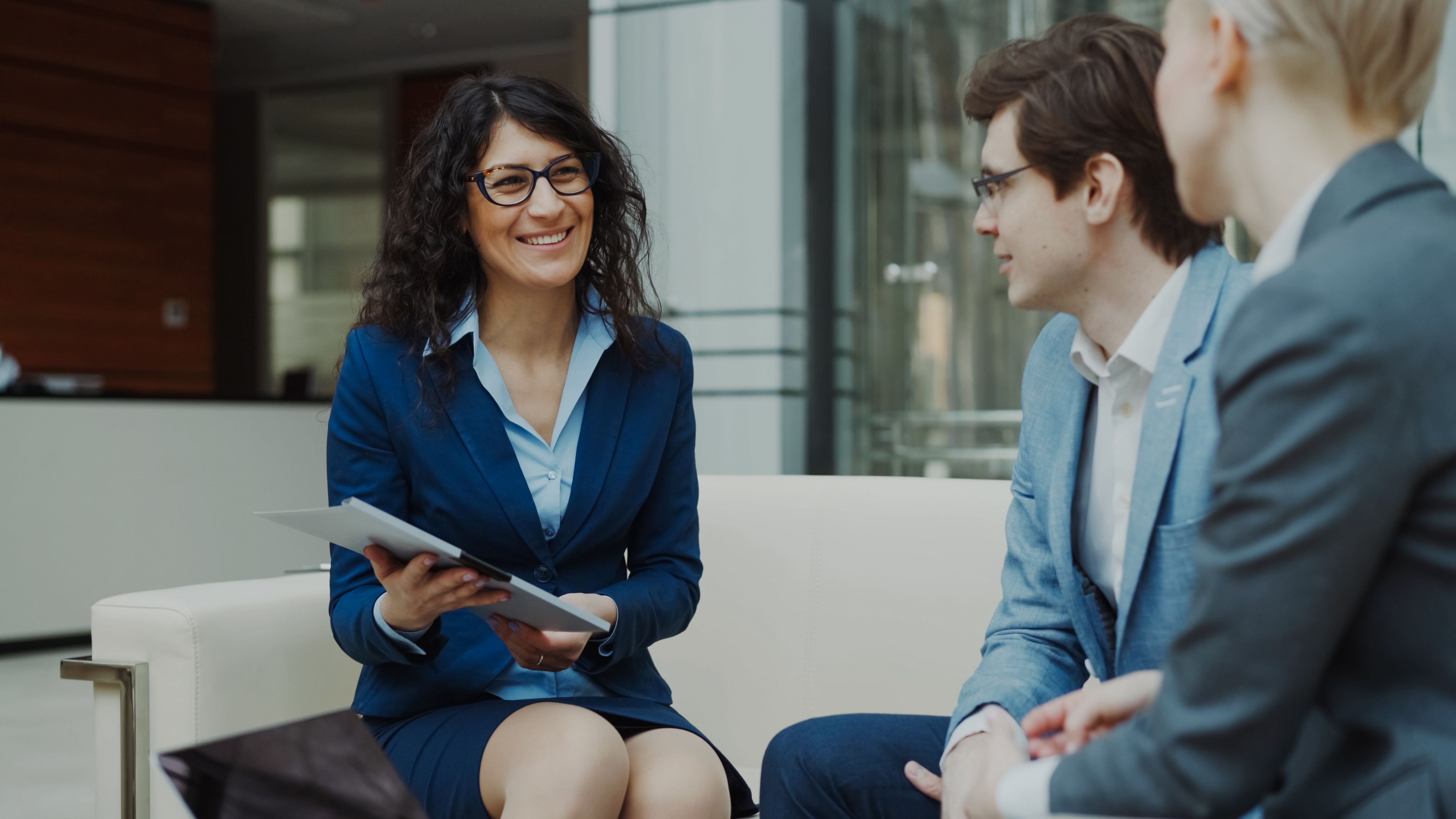 A female advisor holding documents and speaking with two clients sitting on a couch in a modern corporate office lobby.