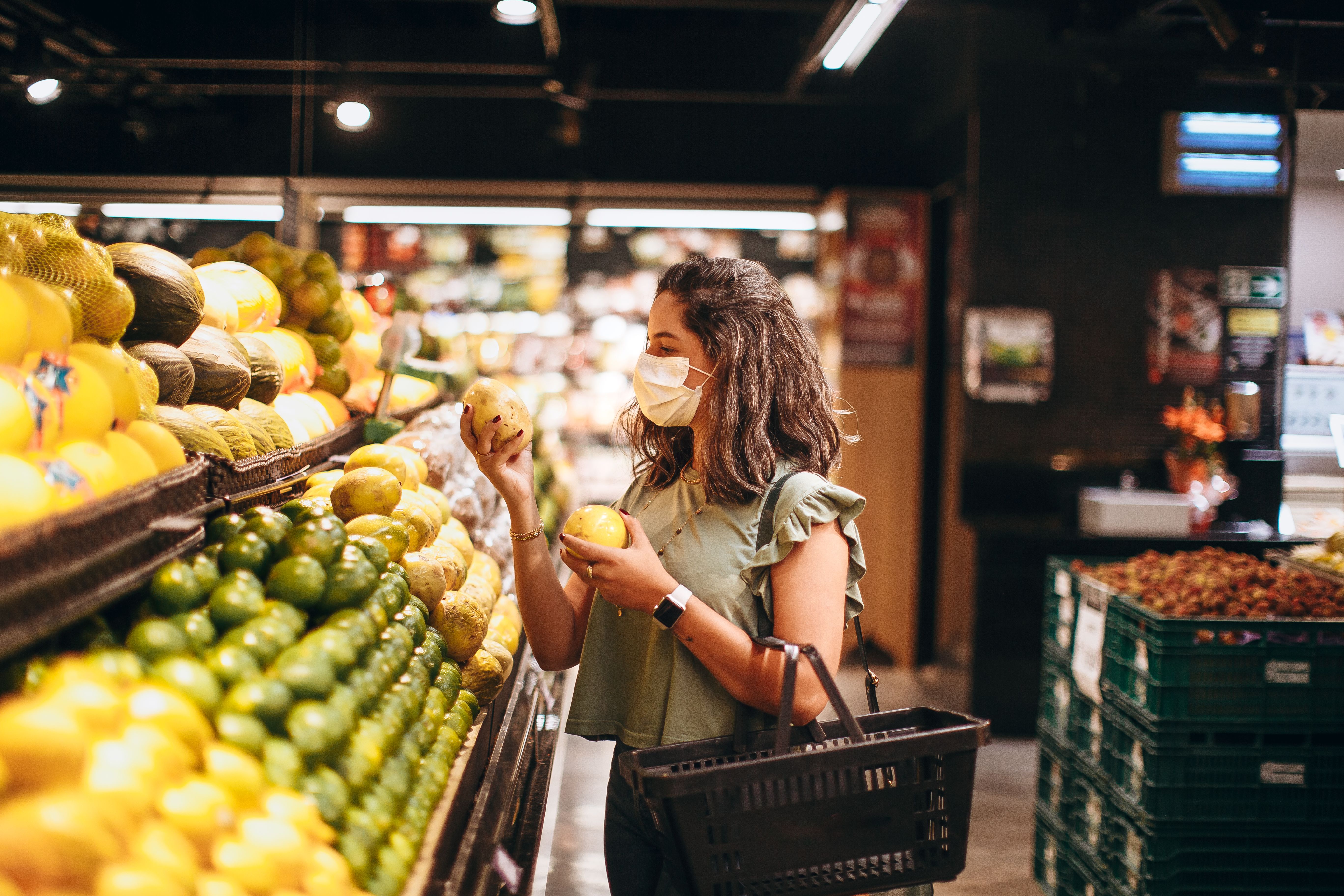 A woman wearing a white face mask selecting fresh yellow produce from the produce section displays at a grocery store.