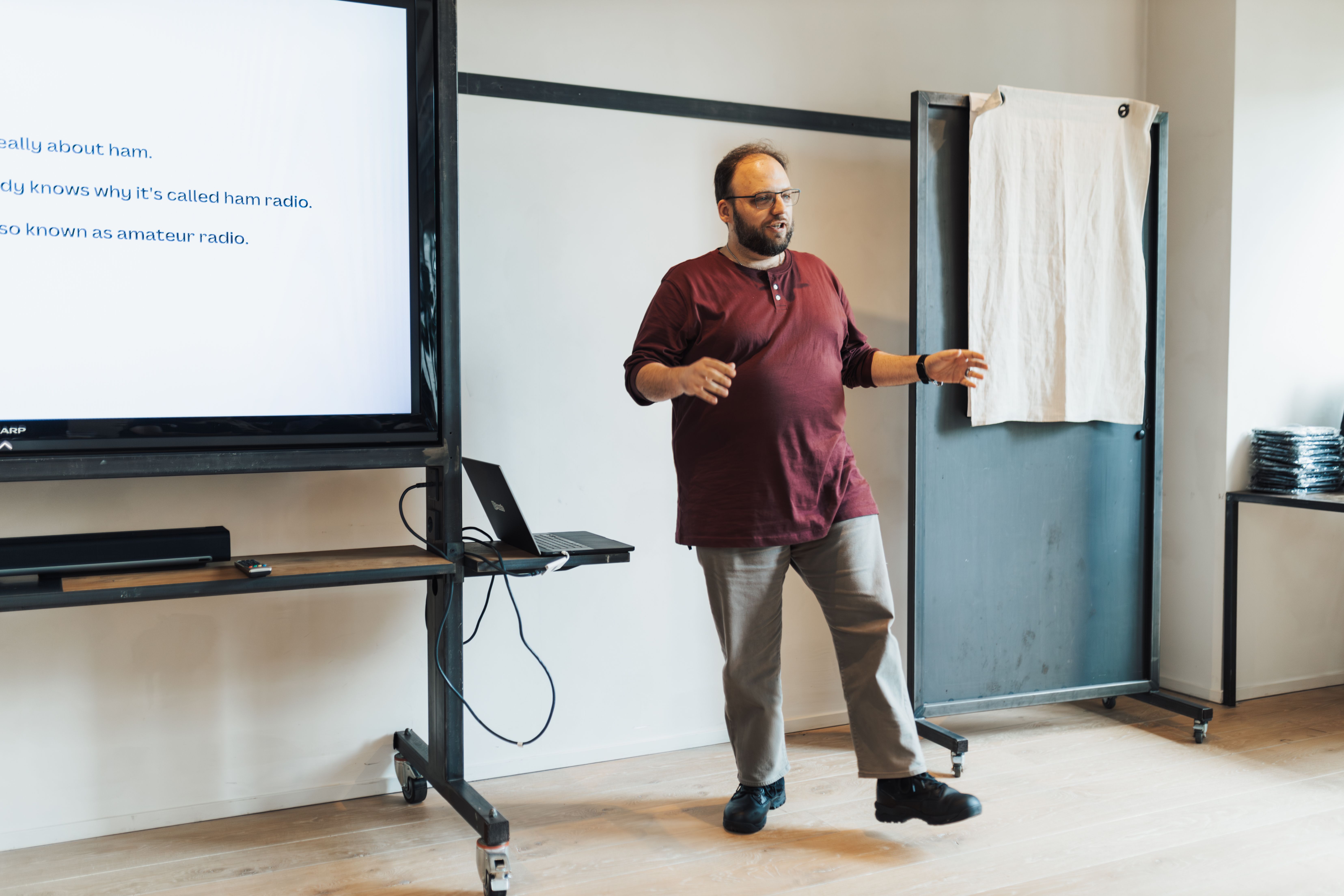 A instructor in a maroon shirt gesturing during a presentation next to a large digital screen display.