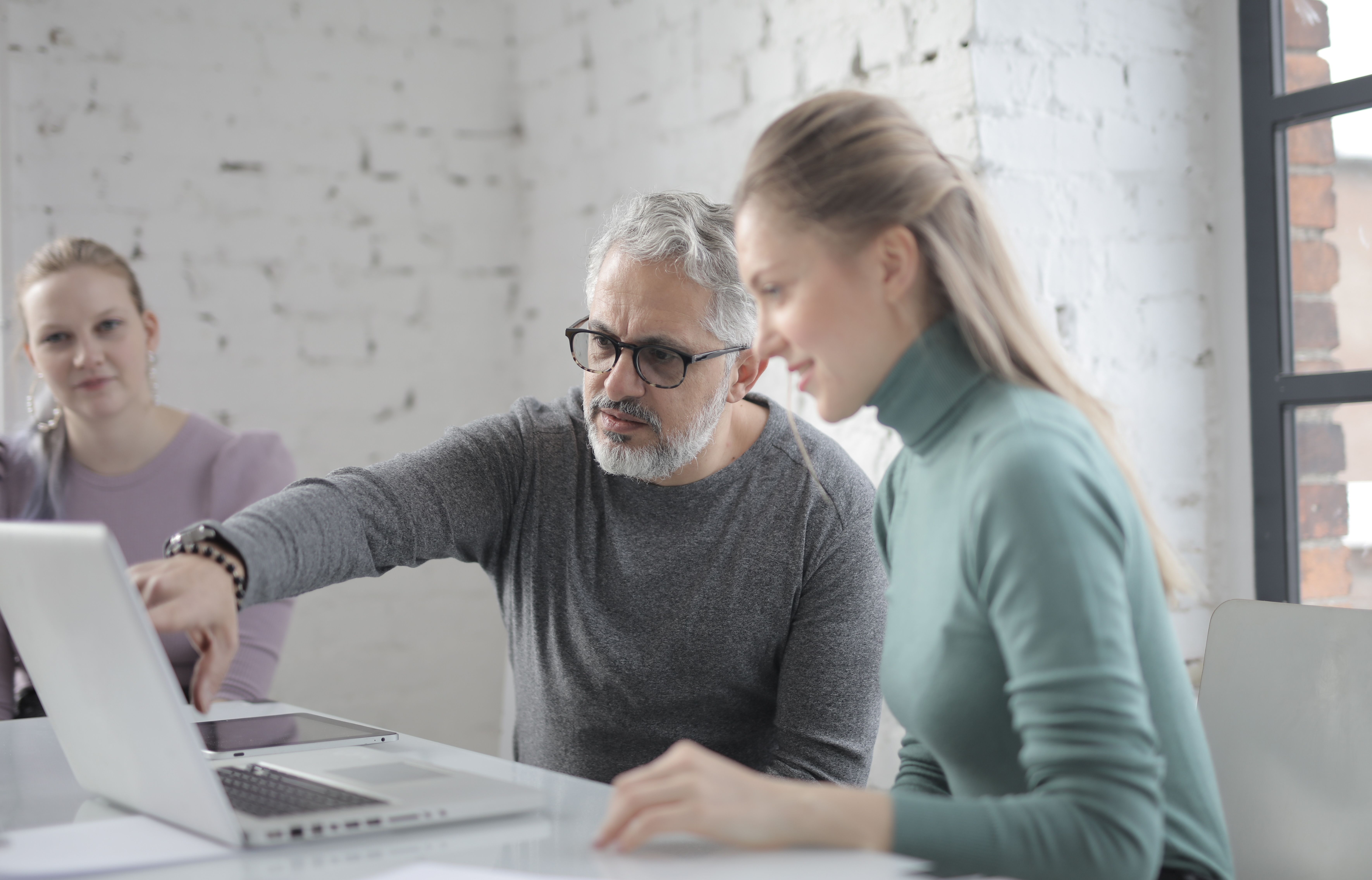 A man with grey hair and glasses pointing at a laptop screen while reviewing work with two female colleagues.