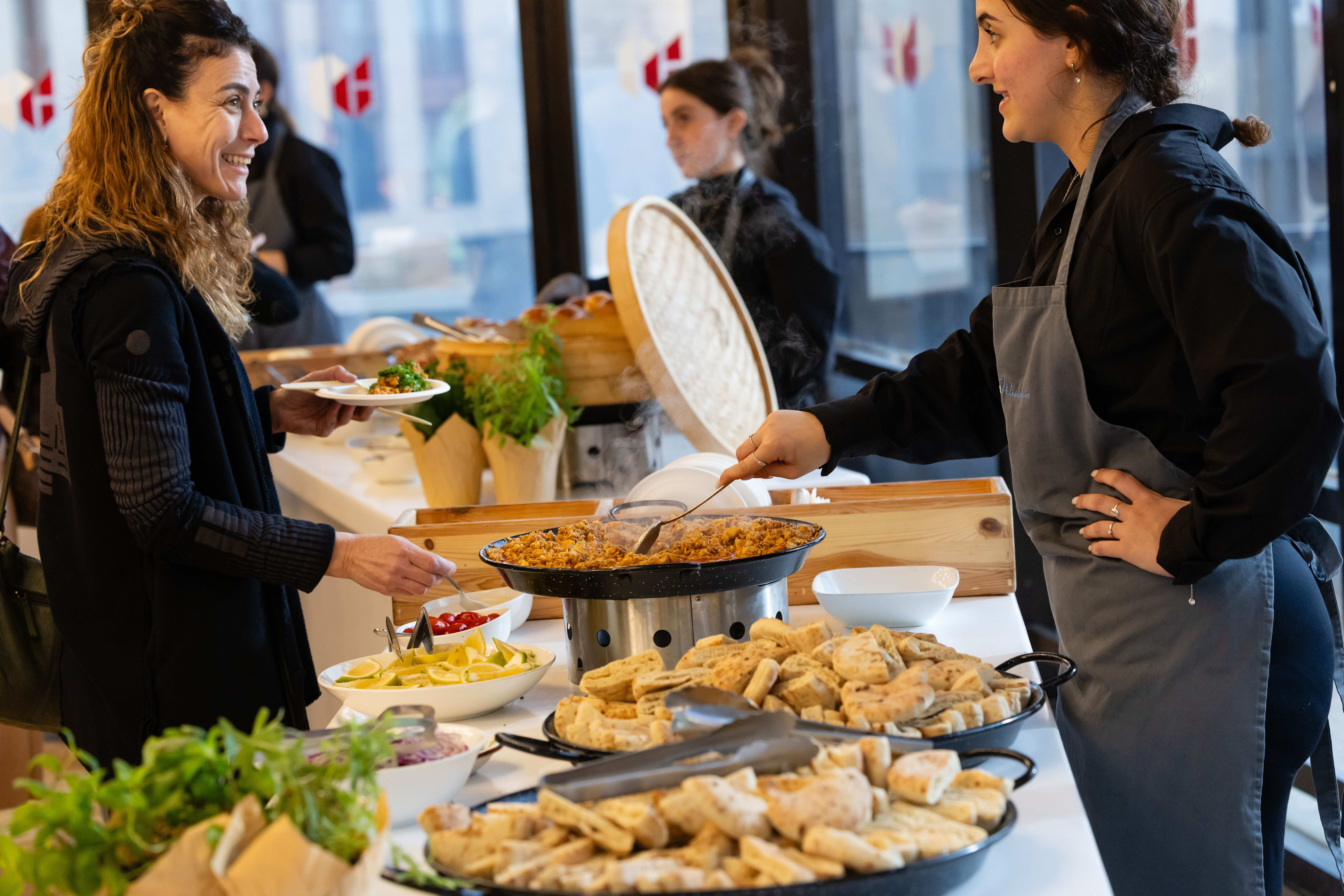 A server in an apron serving hot paella from a large pan to a smiling guest at a buffet station.