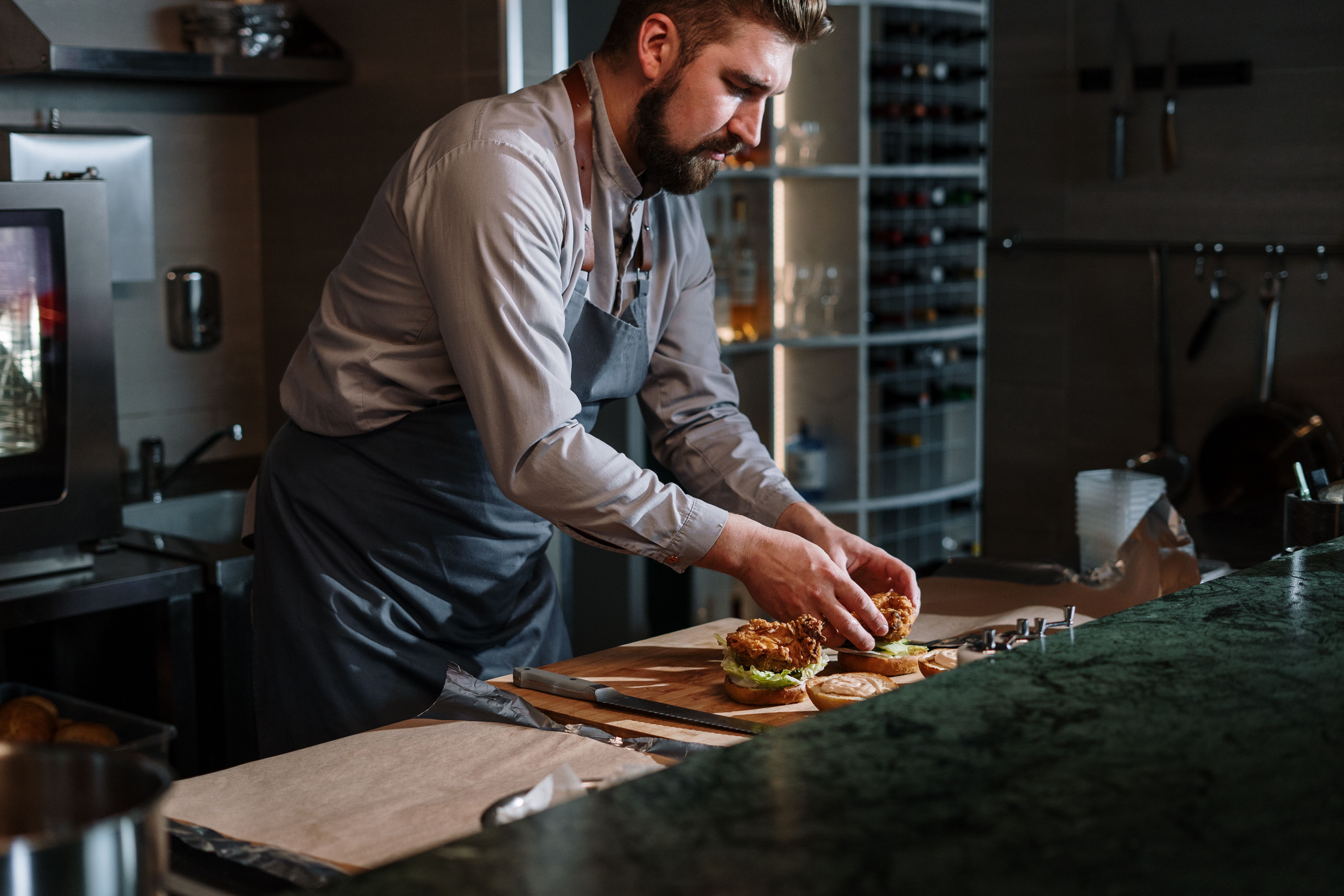 A male chef wearing an apron carefully assembling gourmet burgers on a wooden prep counter in a commercial kitchen.