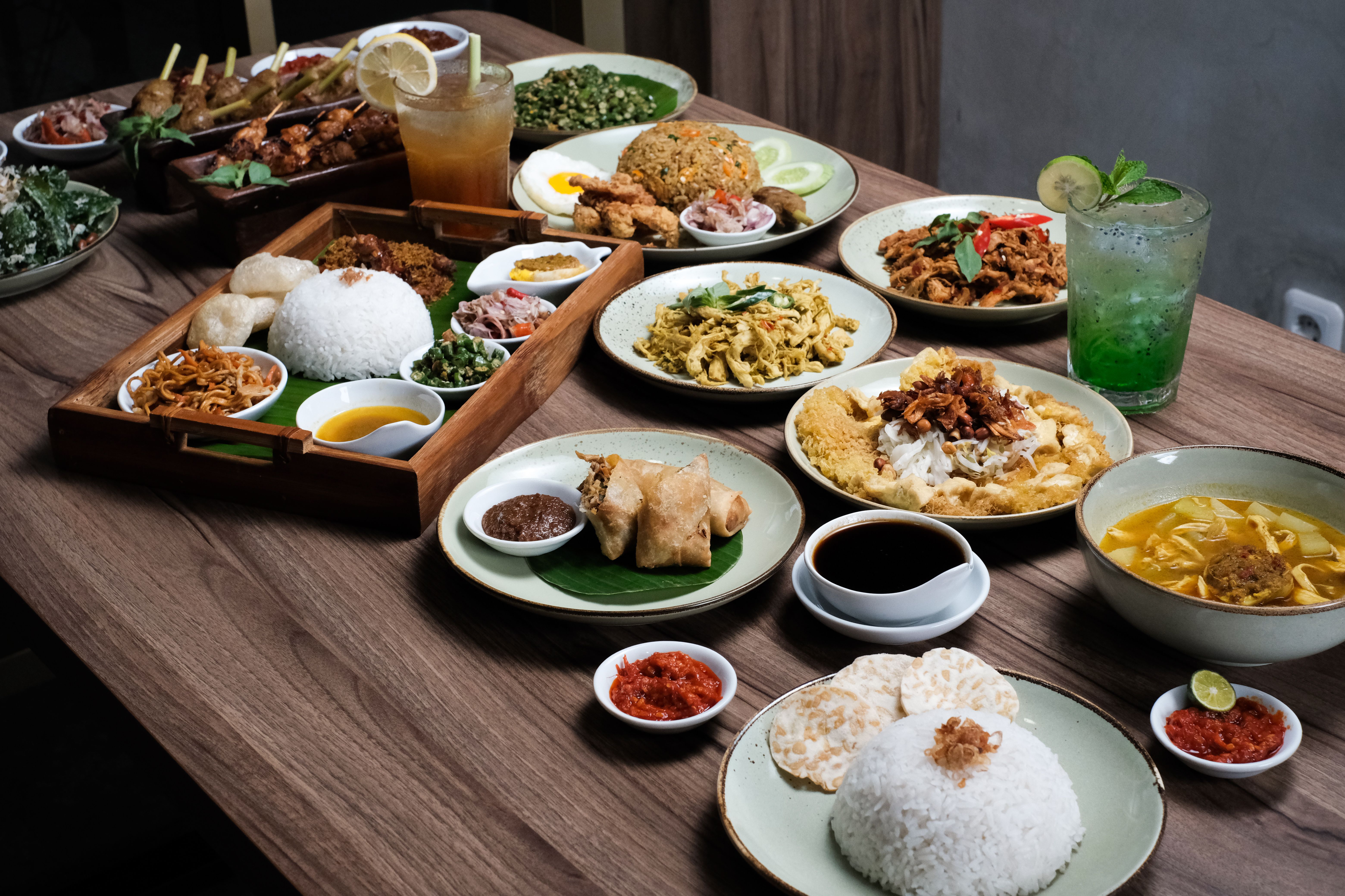 A wooden dining table filled with a large spread of traditional Asian dishes, white rice, skewers, and cold beverages.