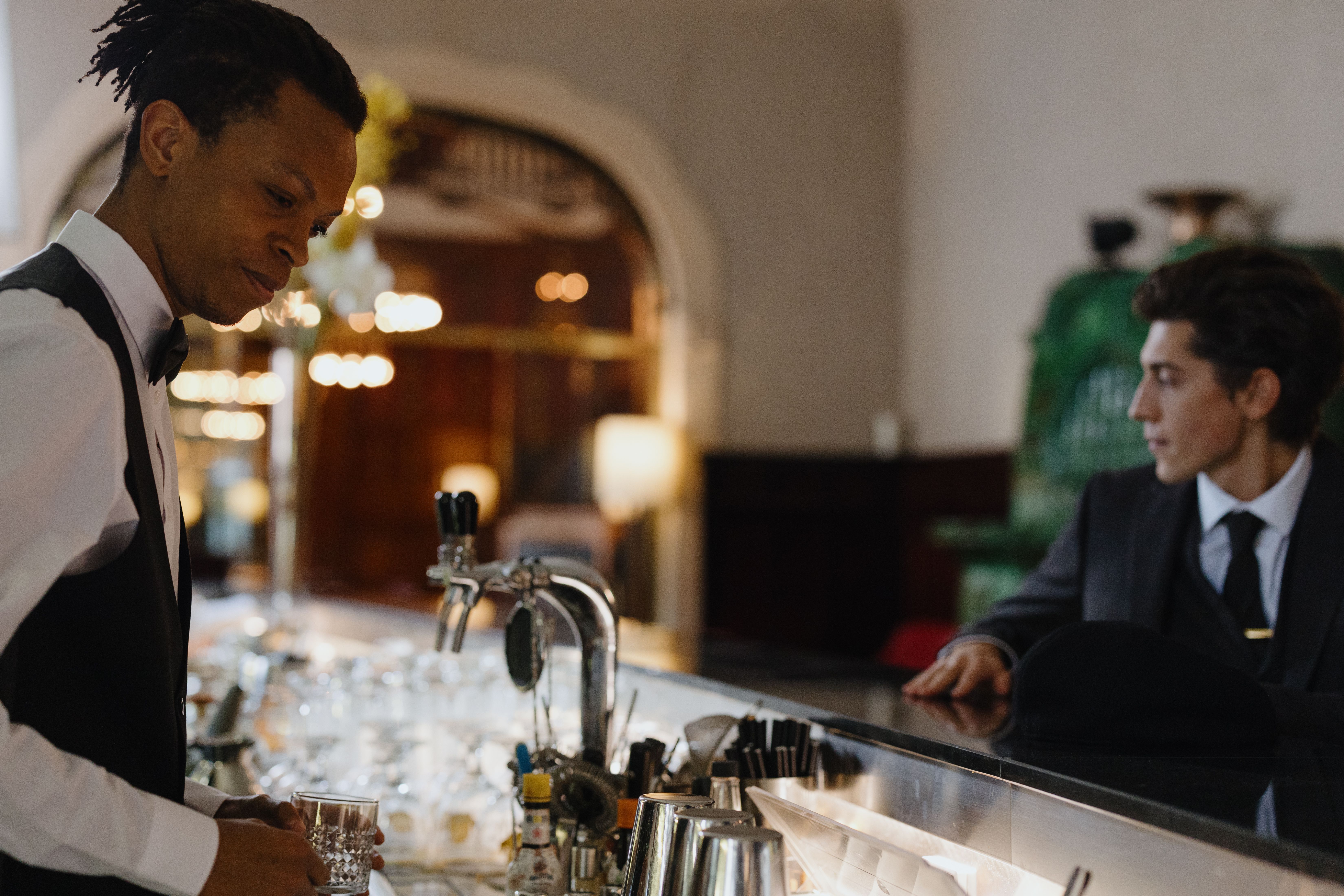 A bartender in a vest and bow tie filling a glass at an upscale bar counter while a seated customer looks on.