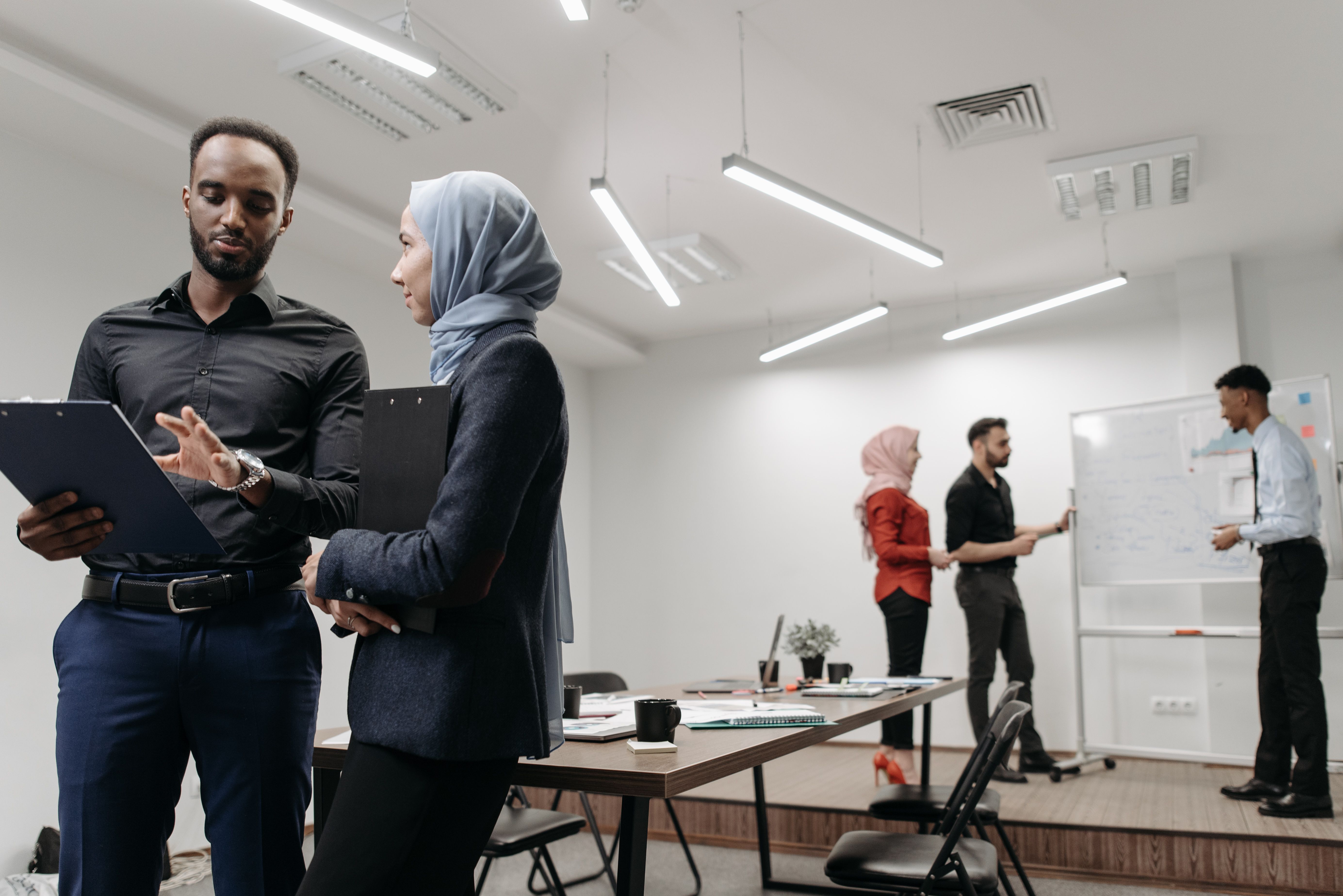Two corporate colleagues holding black clipboards and reviewing notes in a modern, well-lit office setting.