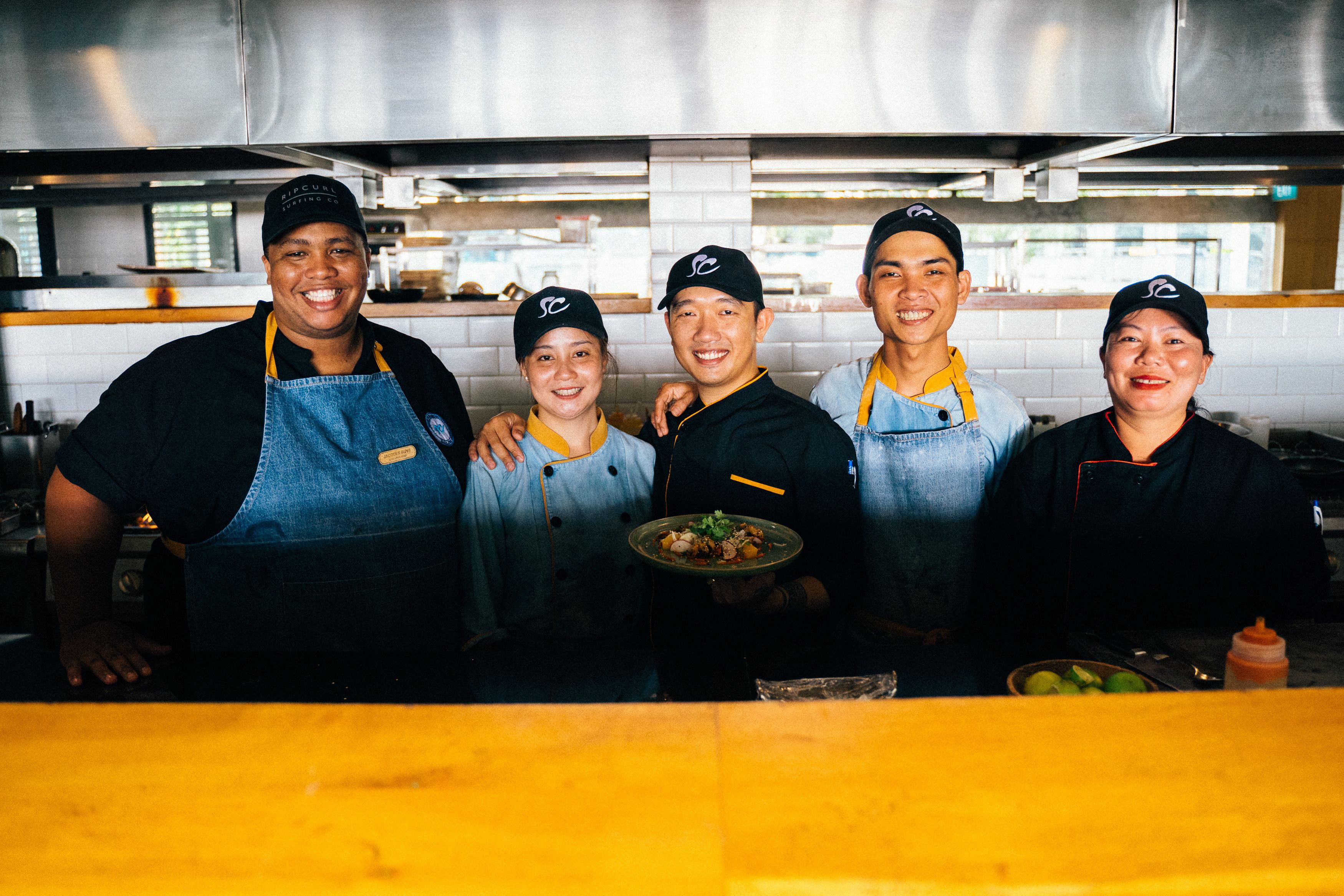A group of five smiling restaurant chefs in matching uniforms and caps standing together in a commercial kitchen.