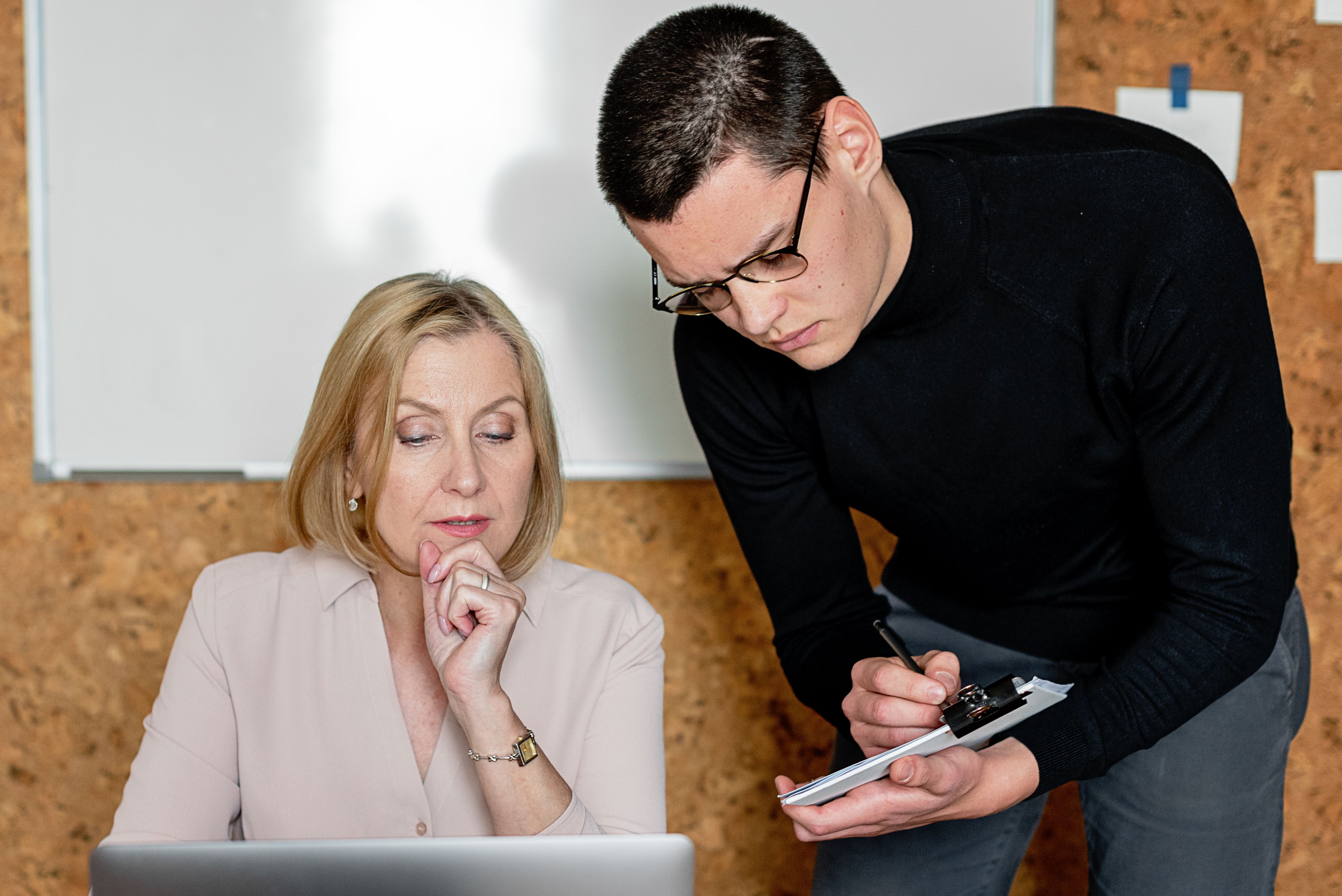 A young man writing on a notepad while reviewing laptop content with a female colleague in an office setting.