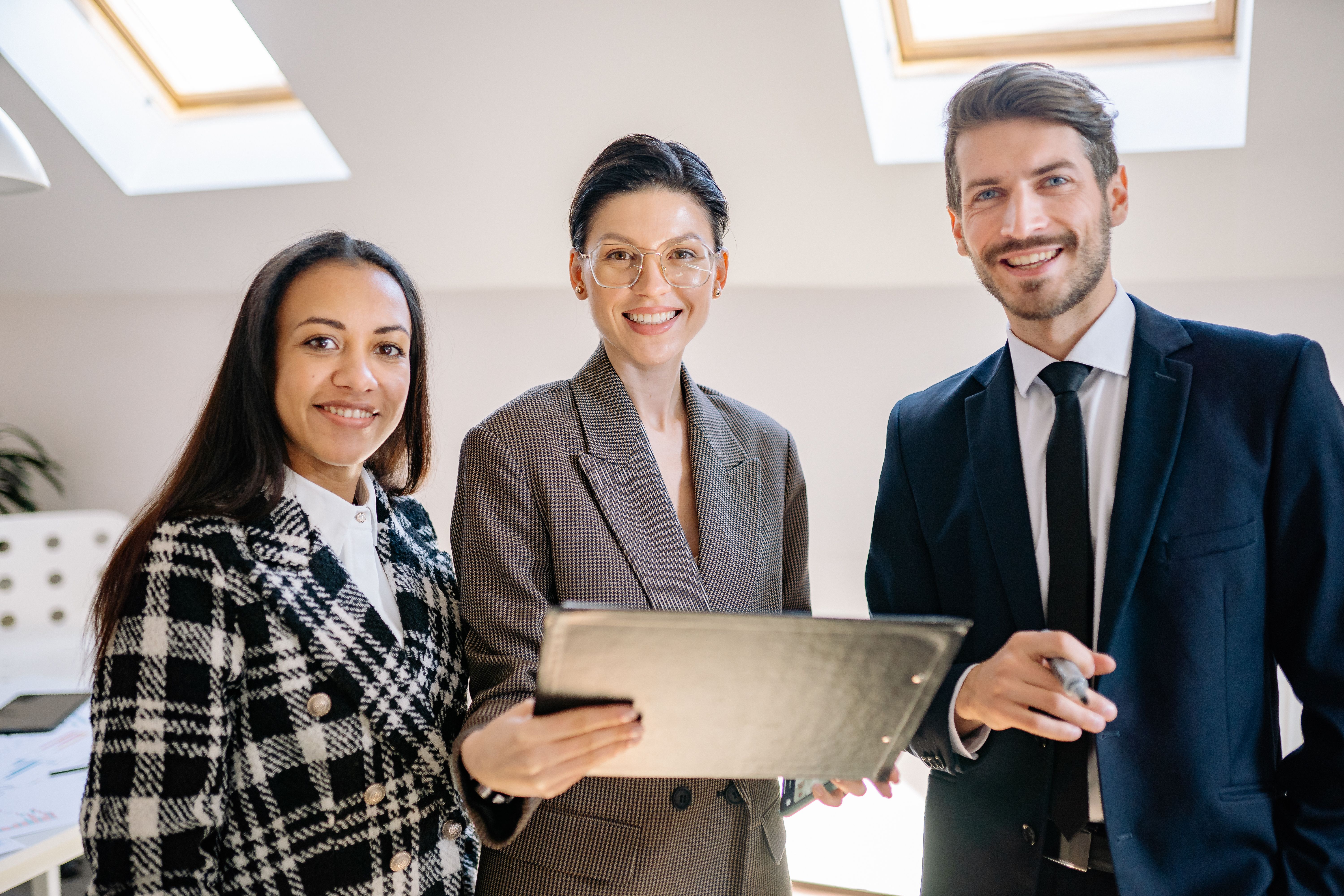 Three smiling professionals in business attire holding a folder and standing together in a modern office space.