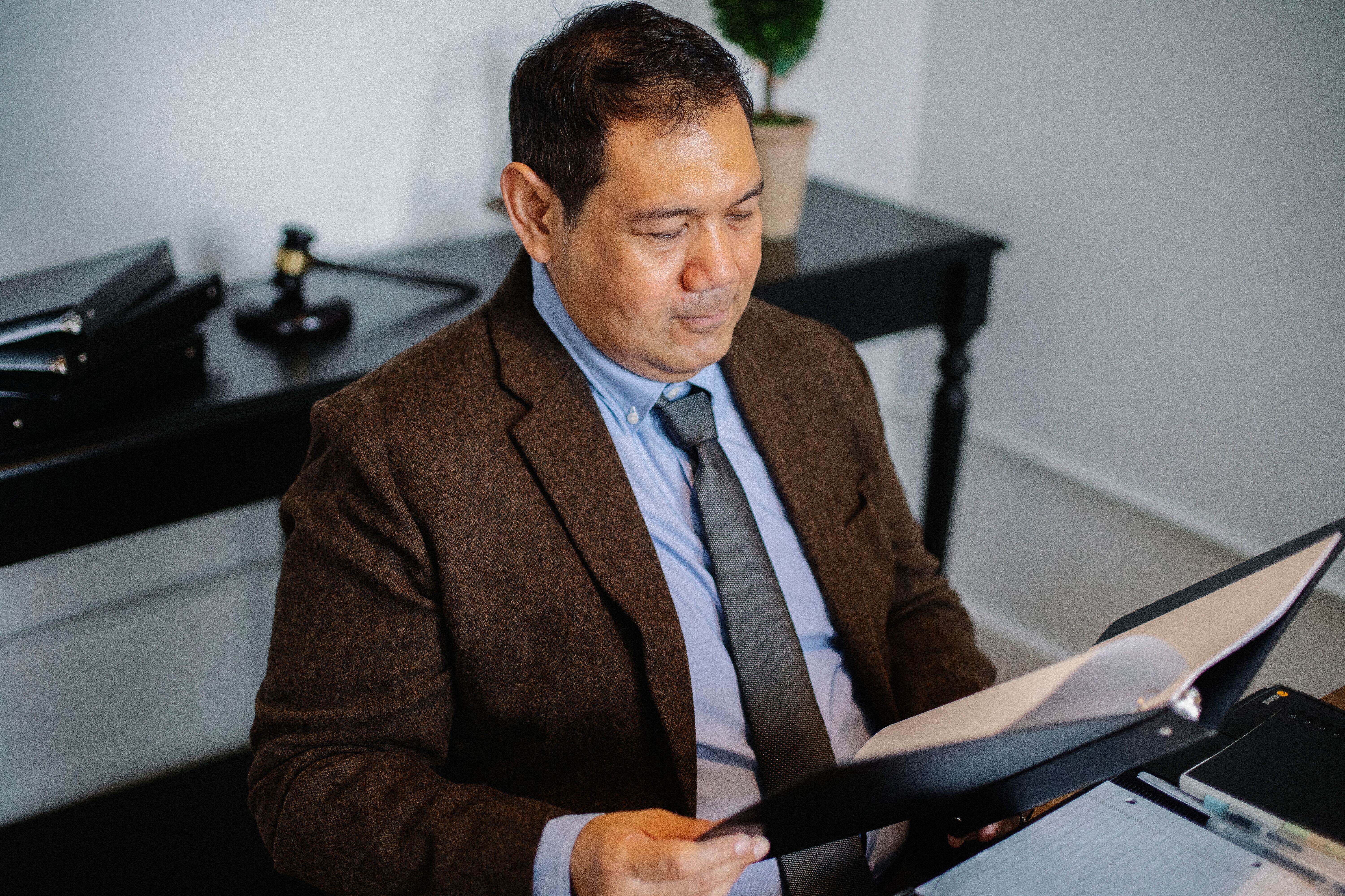 A man in a suit jacket and tie reading through documents held in a black binder at a desk