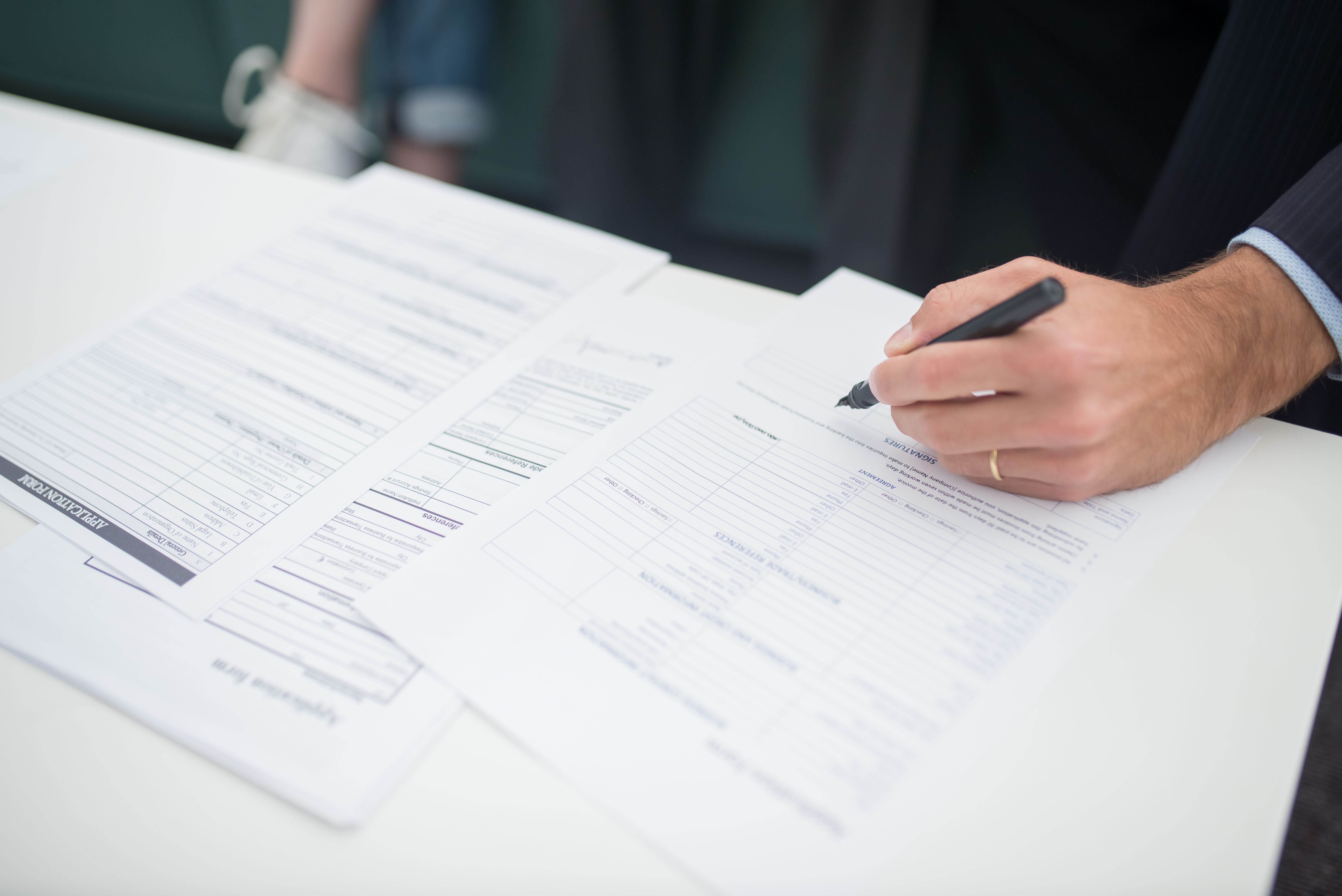 Close-up of a person's hand holding a pen and filling out official application forms on a white table.